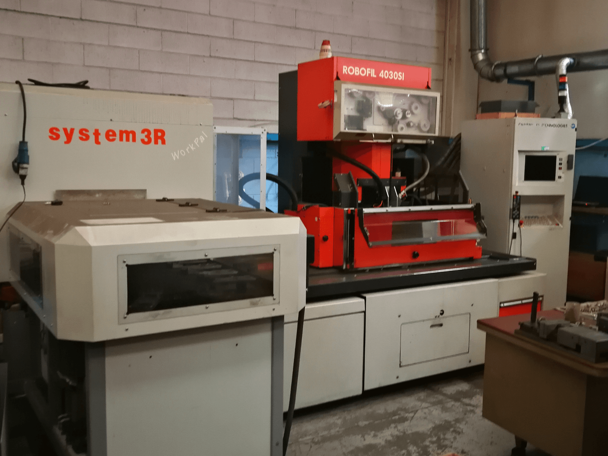 Vue avant de la machine d'électroérosion à fil CHARMILLES ROBOFIL 4030 Si, avec un extérieur rouge et gris et un poste de travail system3R adjacent.
