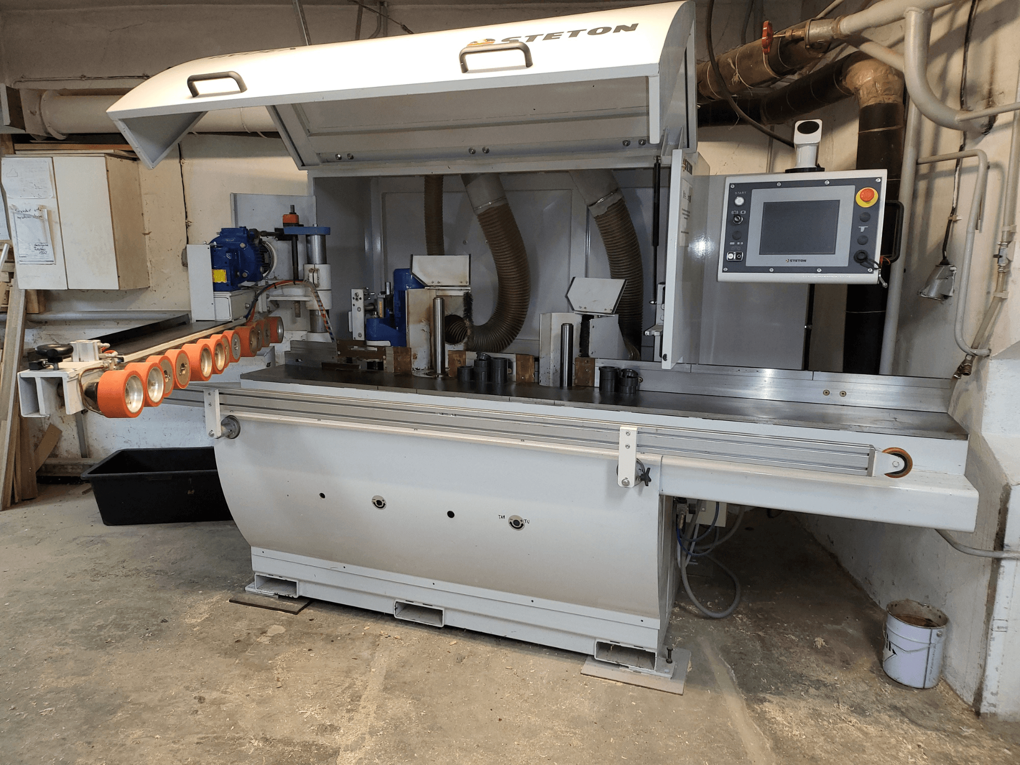 Vue de face d'une machine à bois Stetton avec un panneau de commande, des tubes d'alimentation et diverses pièces de bois sur la table.
