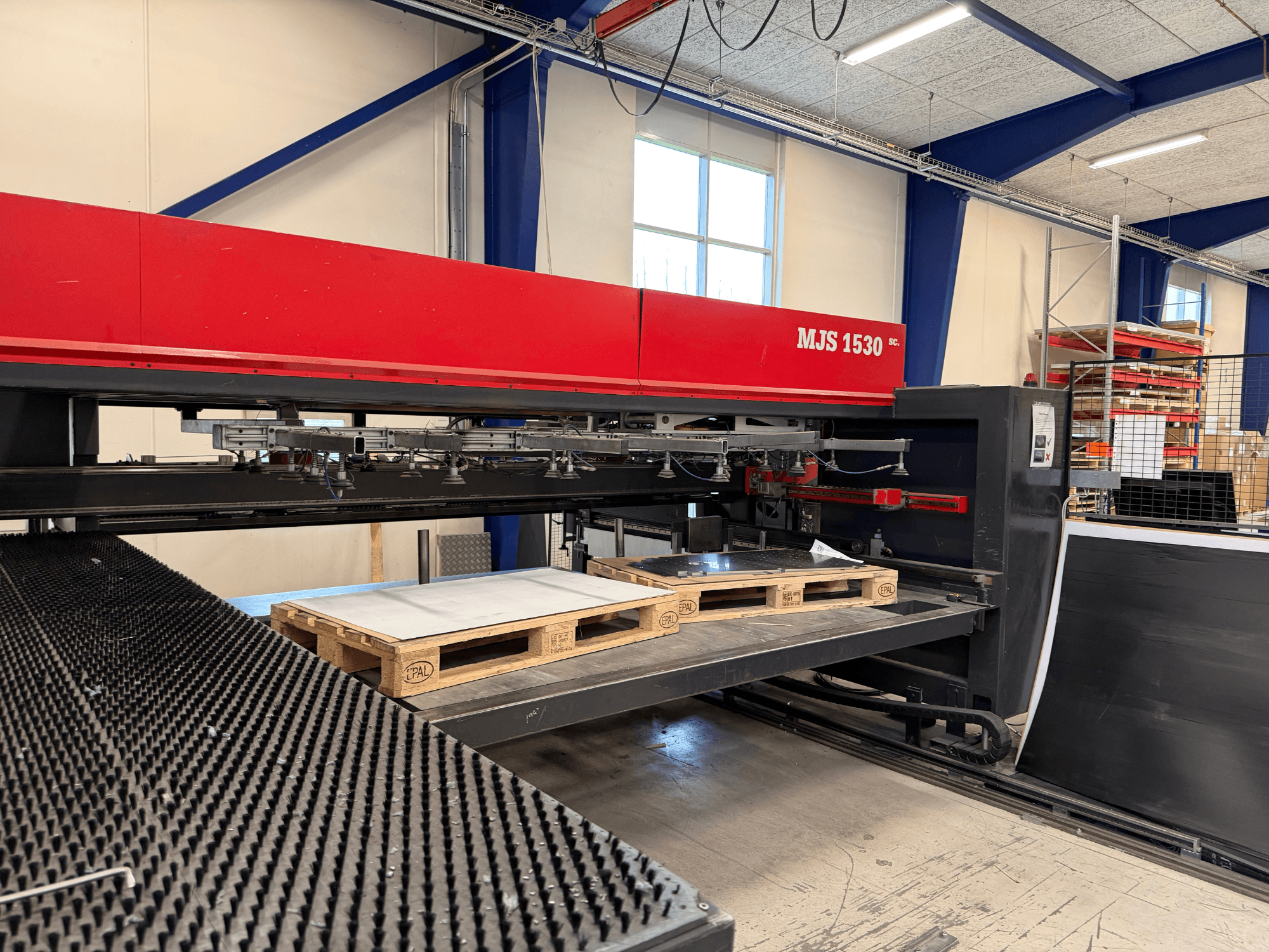 Machine CNC MJS 1530 dans un atelier, vue de face, avec une palette en bois contenant une feuille de métal sur une plate-forme surélevée.