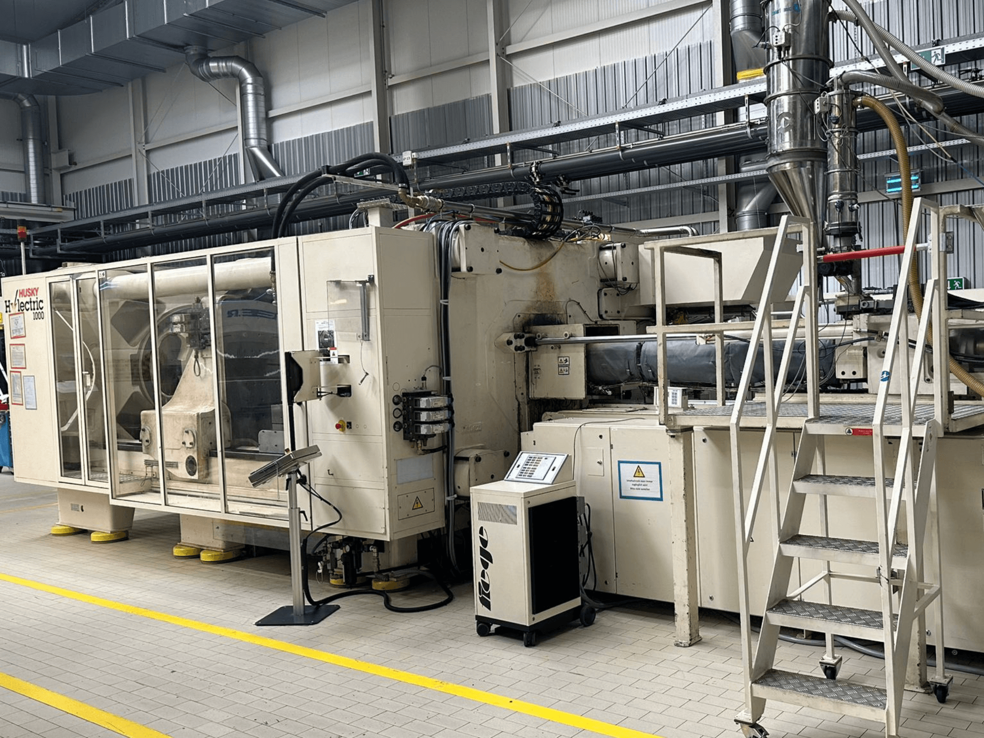 Vue de face d'une presse à injection électrique Husky Hylelectric H1000 RS95/80 dans un environnement industriel, avec son panneau de commande et ses marches.