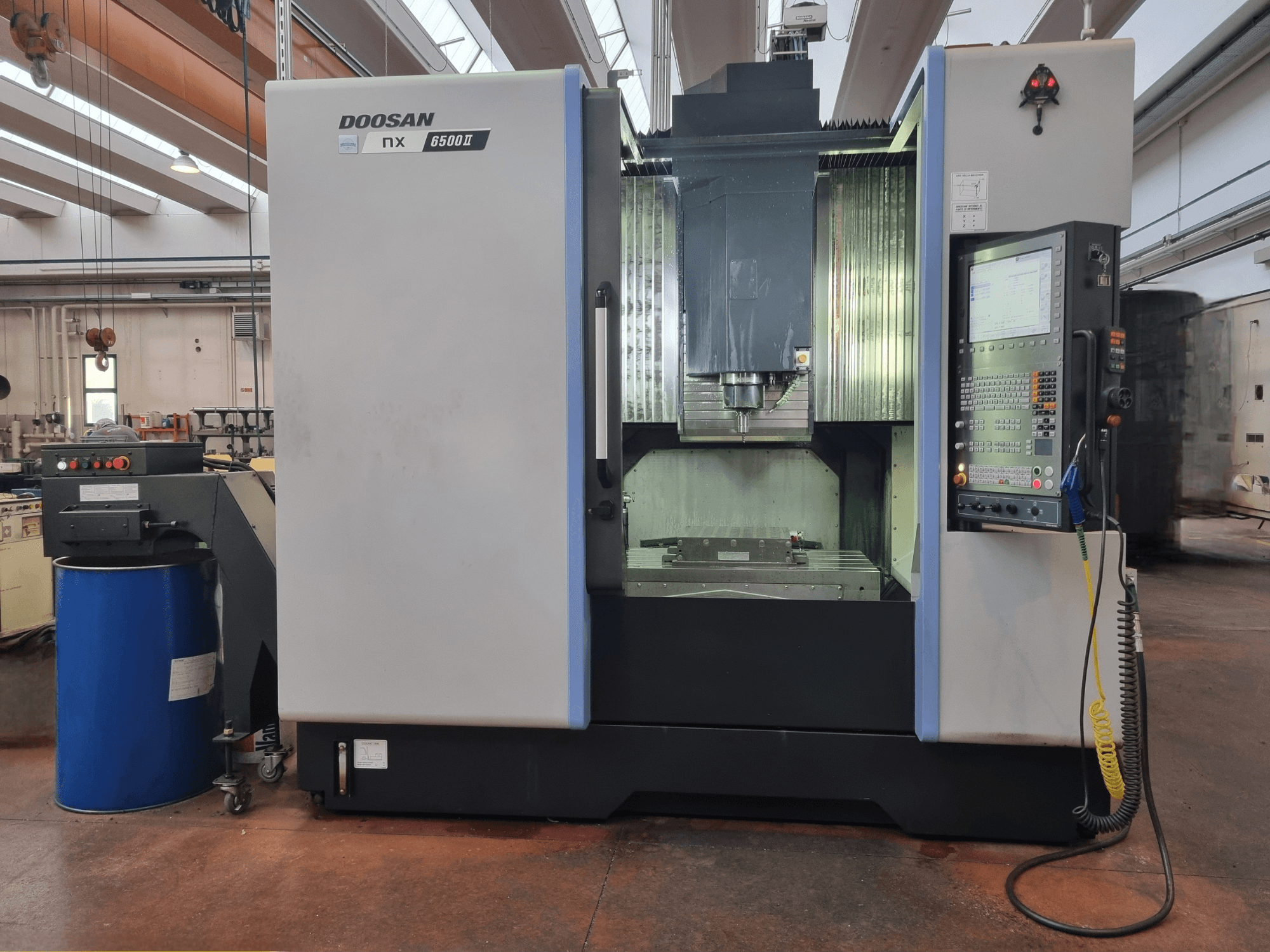 Machine CNC Doosan NX 6500 II, vue de face, avec panneau de commande et zone d'usinage, placée à côté d'un tonneau bleu.
