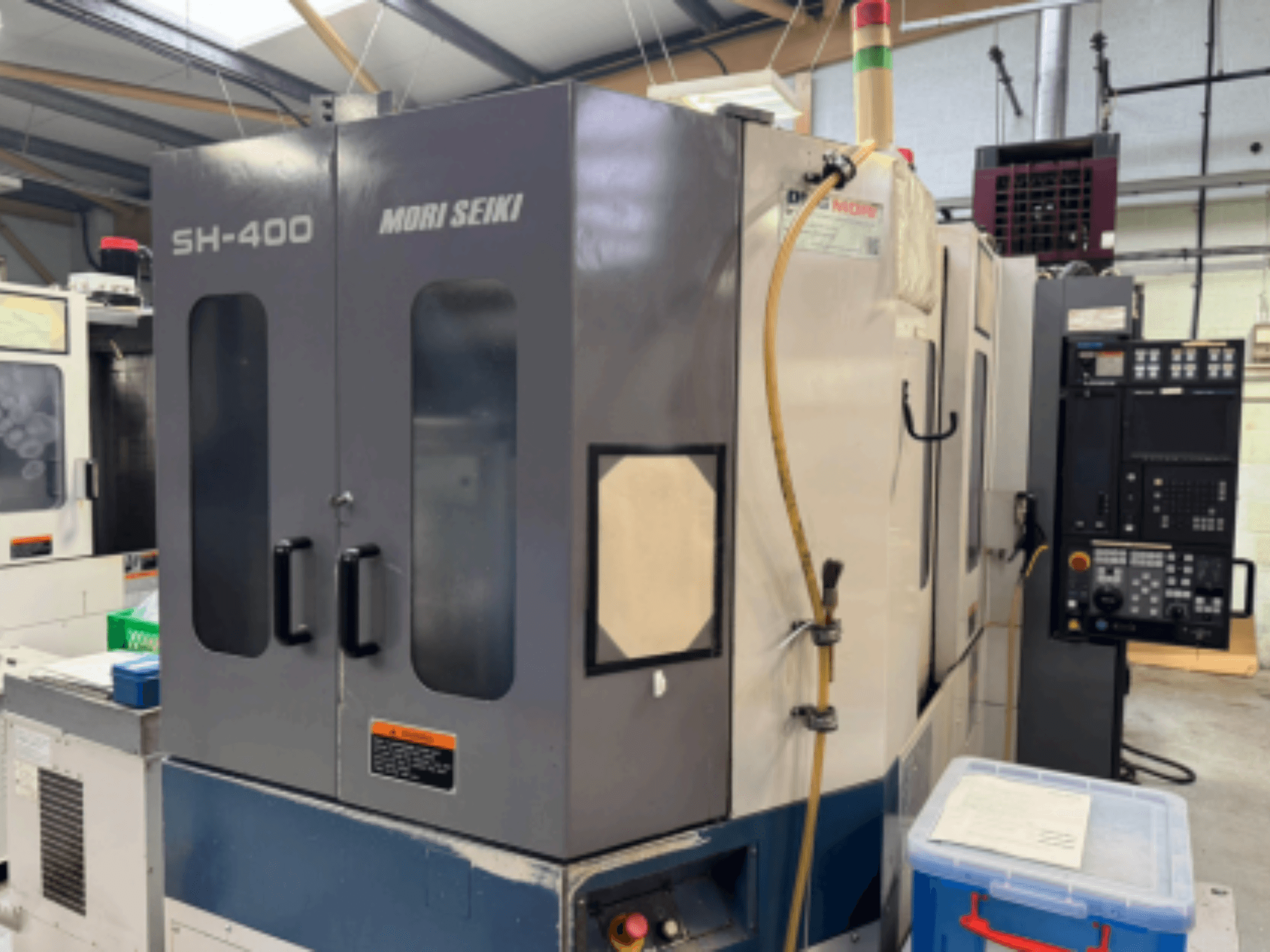 Machine CNC Mori Seiki SH-400, vue de face, avec deux portes, un panneau de commande et un tuyau de fixation. Conteneur de stockage bleu à proximité.
