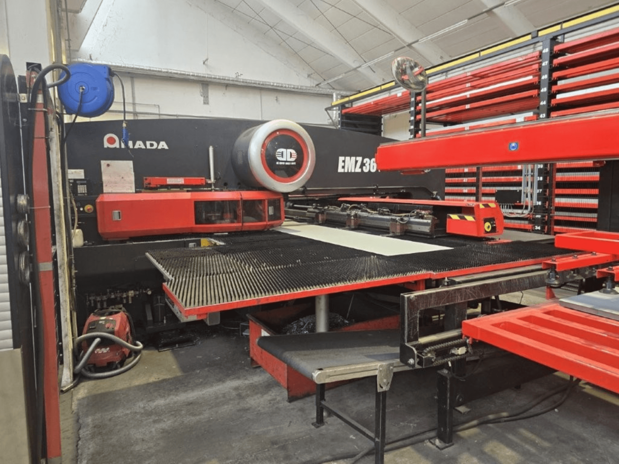 Machine de découpe laser Amada EMZ 36 dans un atelier, vue de face, avec des accents rouges et une grande table de découpe.