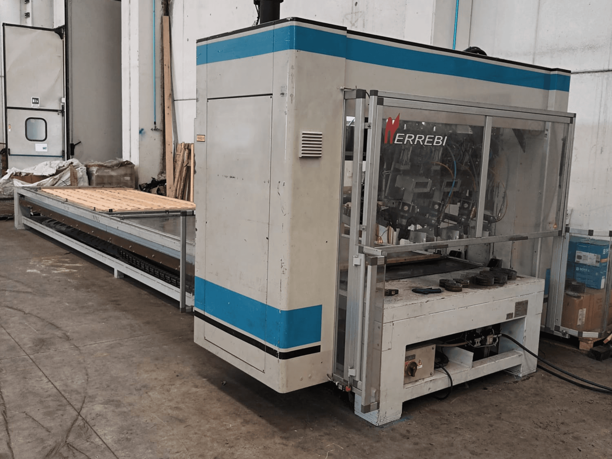 La machine ERREBI, vue de côté, comporte une grande table de traitement avec des composants mécaniques et un couvercle de protection transparent.