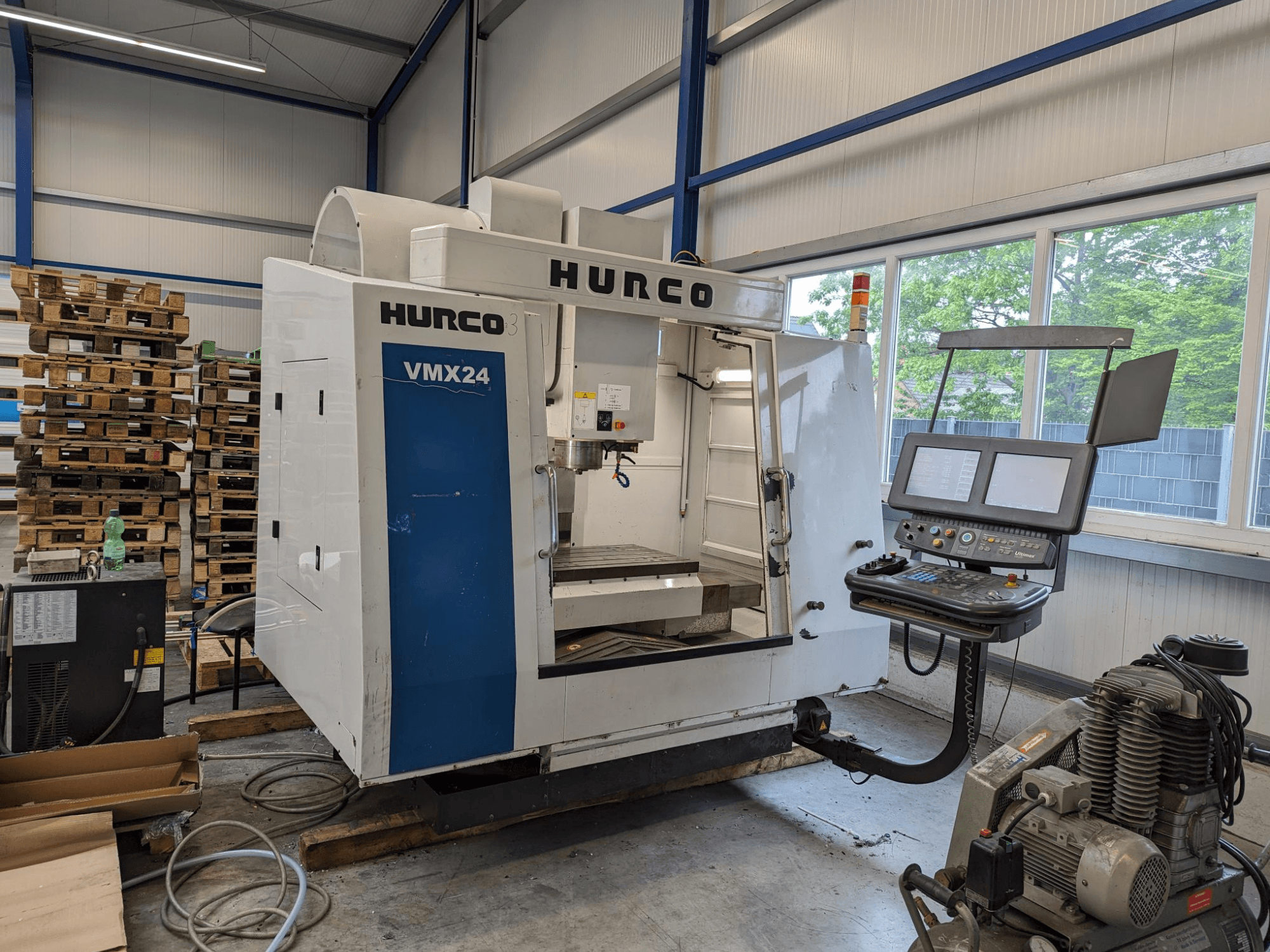Fraiseuse Hurco VMX24, vue de face, avec panneau de commande et zone d'outils ; palettes empilées en arrière-plan ; espace de travail industriel.