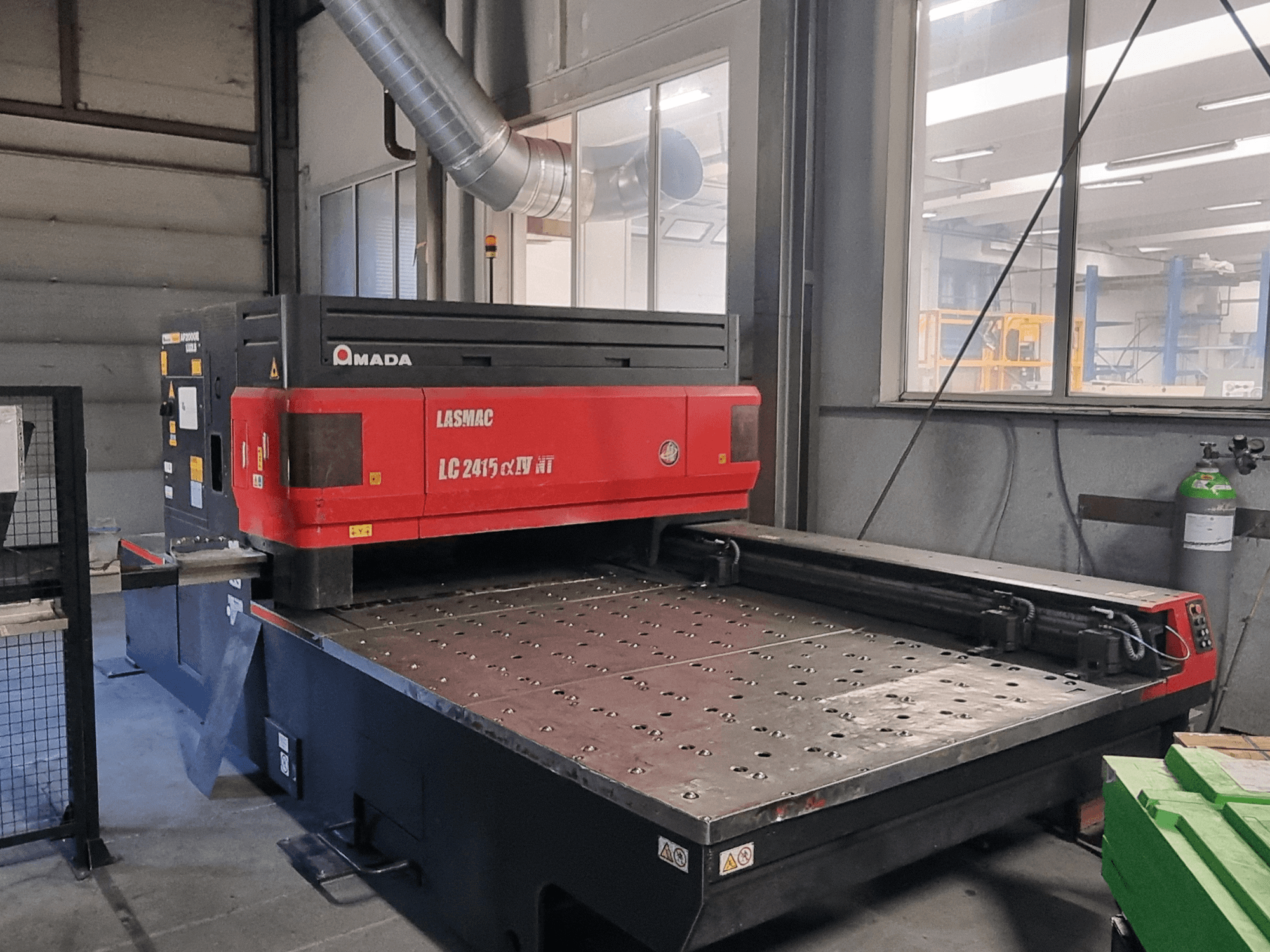 Machine de découpe laser Amada LASMAC LC 2415 αⅡ NT en rouge et noir, vue de face, avec un banc de découpe motorisé et des commandes de précision.