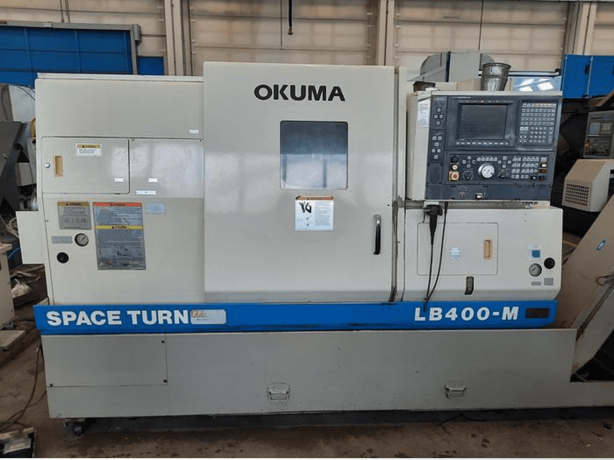 Tour CNC Okuma LB400-M, vue de face avec panneau de commande, étiquettes d'avertissement et marquage bleu "SPACE TURN" visible.