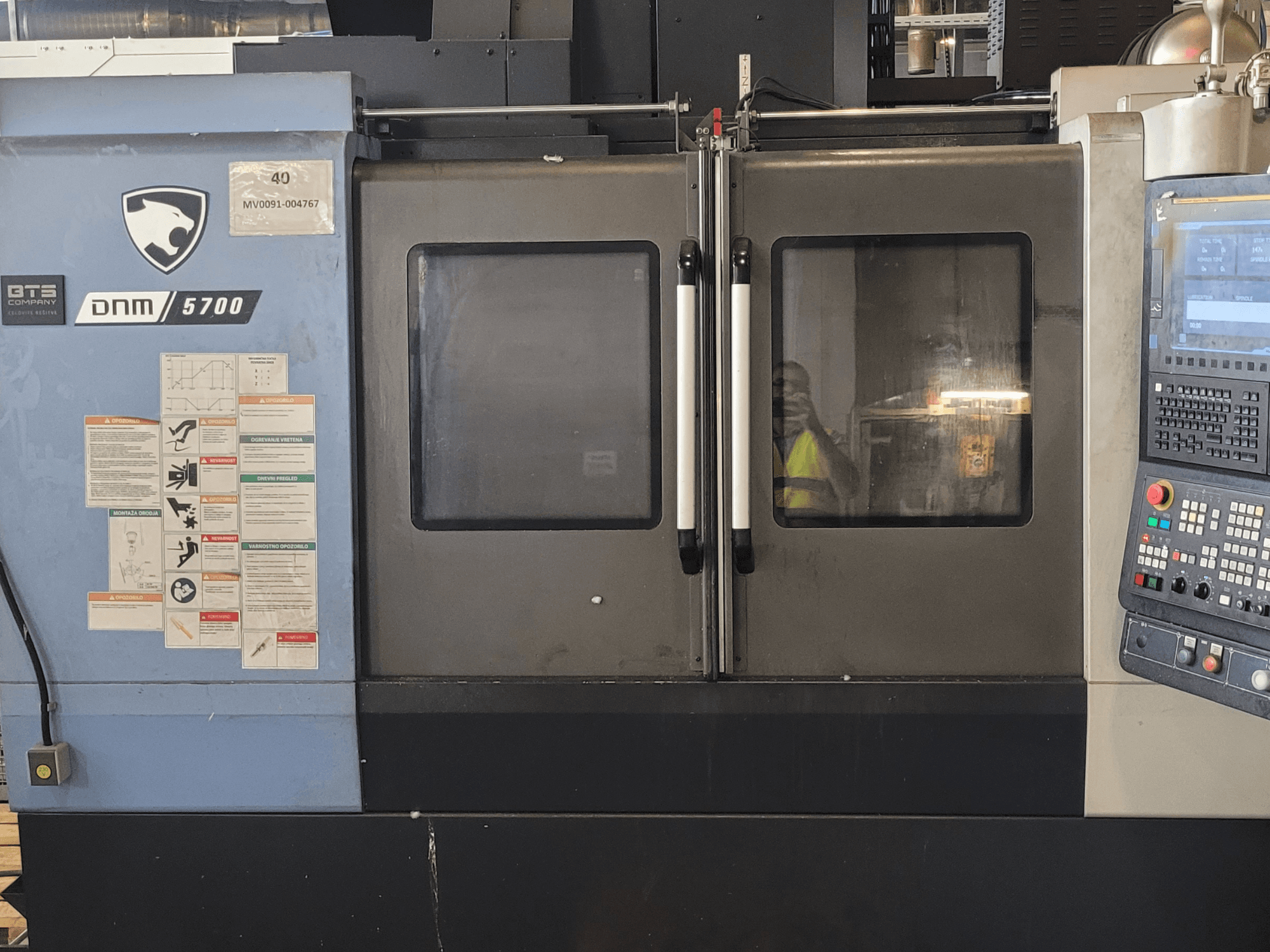Vue de face du centre d'usinage vertical DOOSAN DNM 5700, avec le panneau de commande, les étiquettes de sécurité et les portes légèrement entrouvertes.