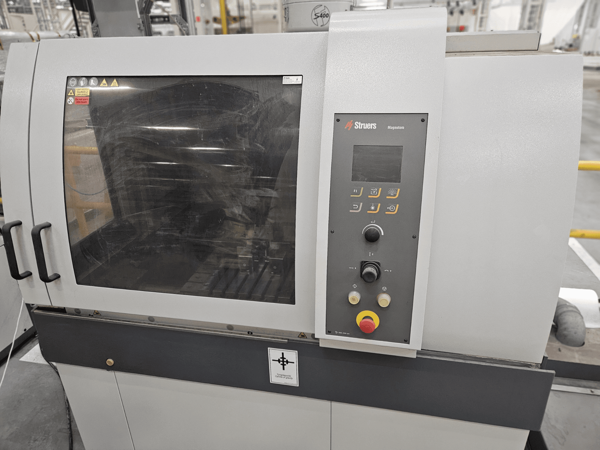 Vue avant de la machine Struers Magpentor avec le panneau de commande, les boutons et une grande fenêtre de visualisation. Les étiquettes techniques sont visibles.