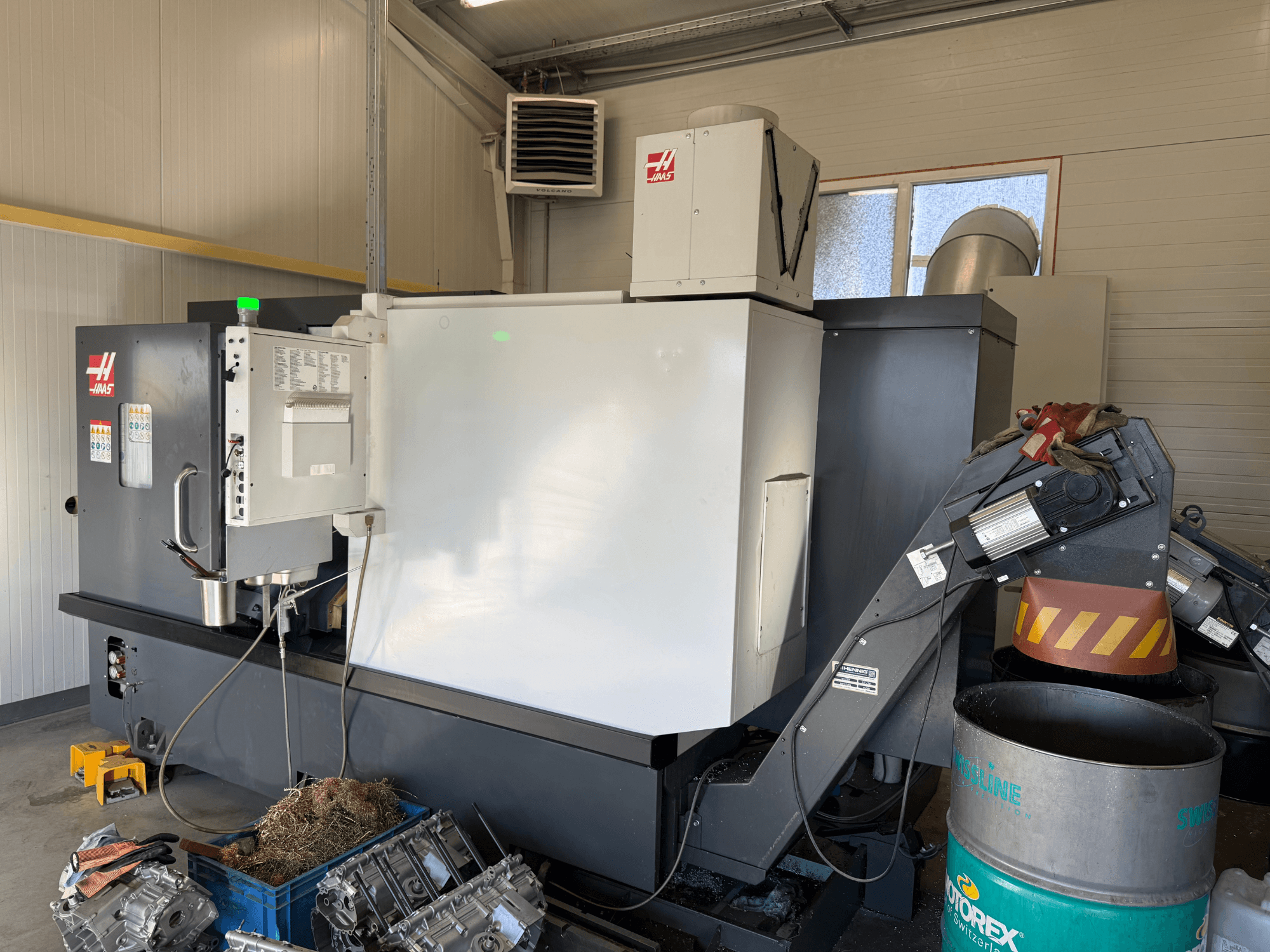 Machine CNC Haas avec panneau de commande, vue latérale ; lignes de refroidissement et d'huile visibles ; déchets métalliques dans une poubelle bleue ; environnement industriel.