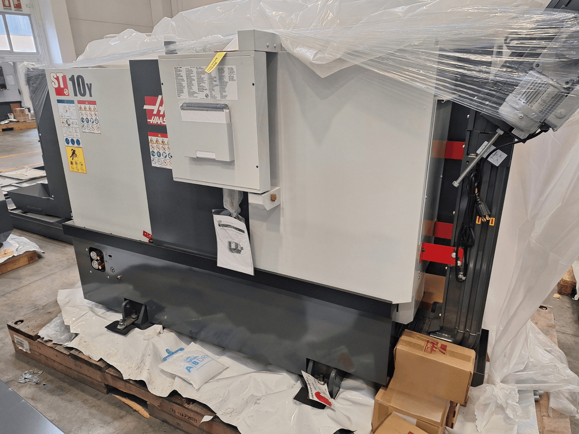 Vue de profil d'un tour horizontal HAAS ST-10Y EU, emballé dans du plastique, avec le panneau de commande et les étiquettes d'avertissement visibles.