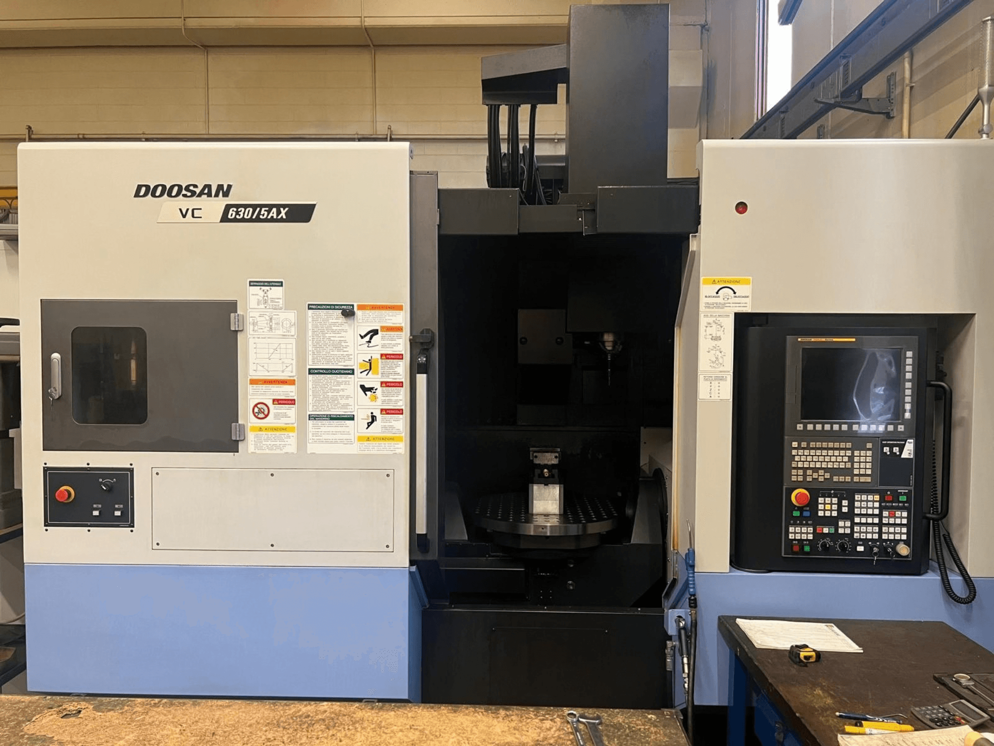 Vue de face d'un centre d'usinage vertical DOOSAN VC 630/5AX, mettant en évidence le panneau de commande et la zone de la broche, avec les étiquettes de sécurité bien visibles.