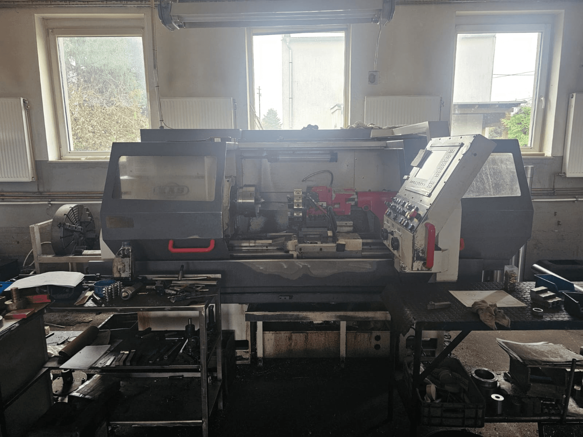 Tour dans un atelier, vue de face, avec panneau de commande et outils sur des tables, entouré de fenêtres et d'un espace de travail encombré.