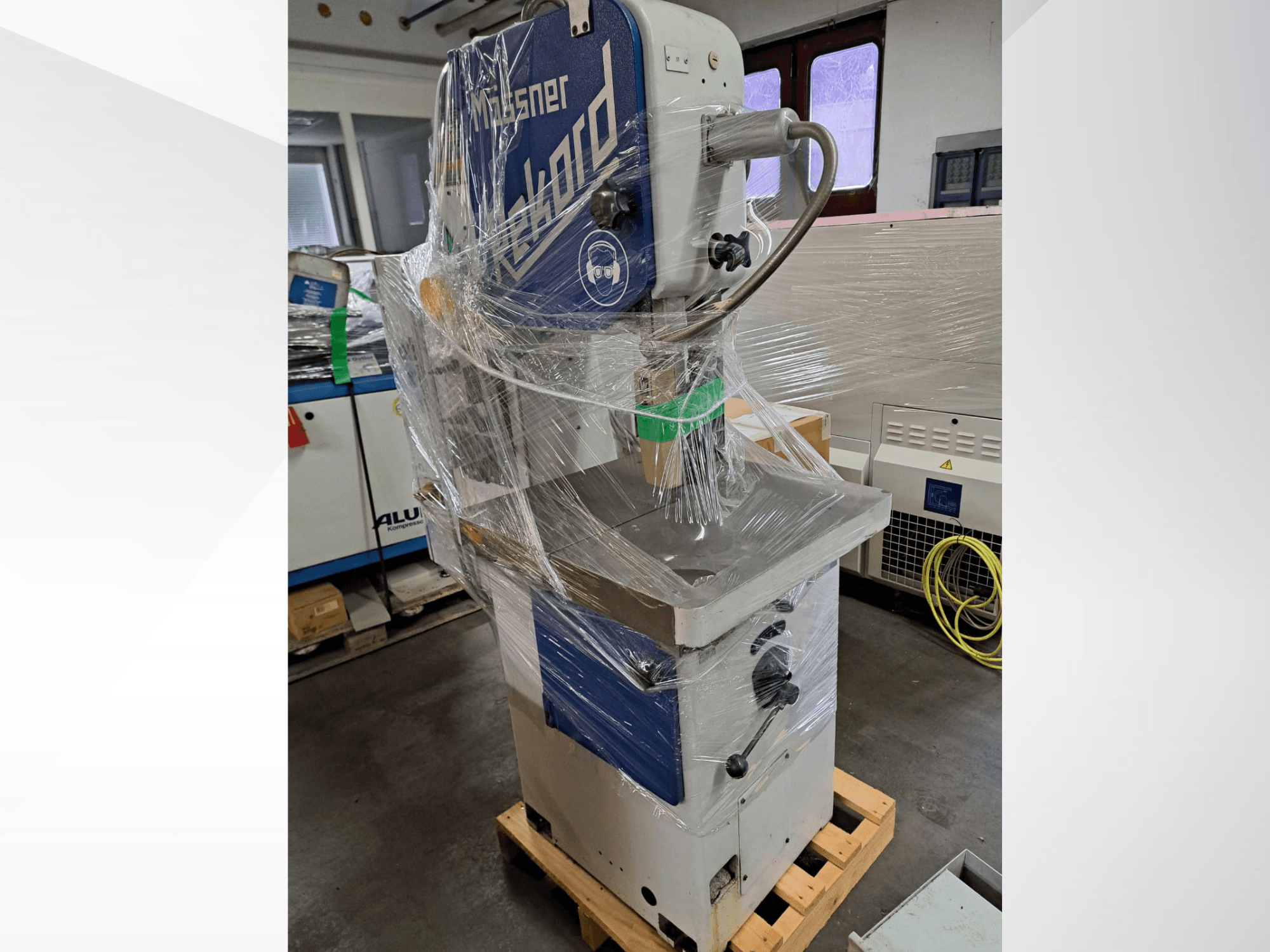 Scie à ruban métallique MOSSNER Rekord emballée dans du plastique ; vue de profil montrant le panneau de commande et le socle posés sur une palette en bois dans un atelier.
