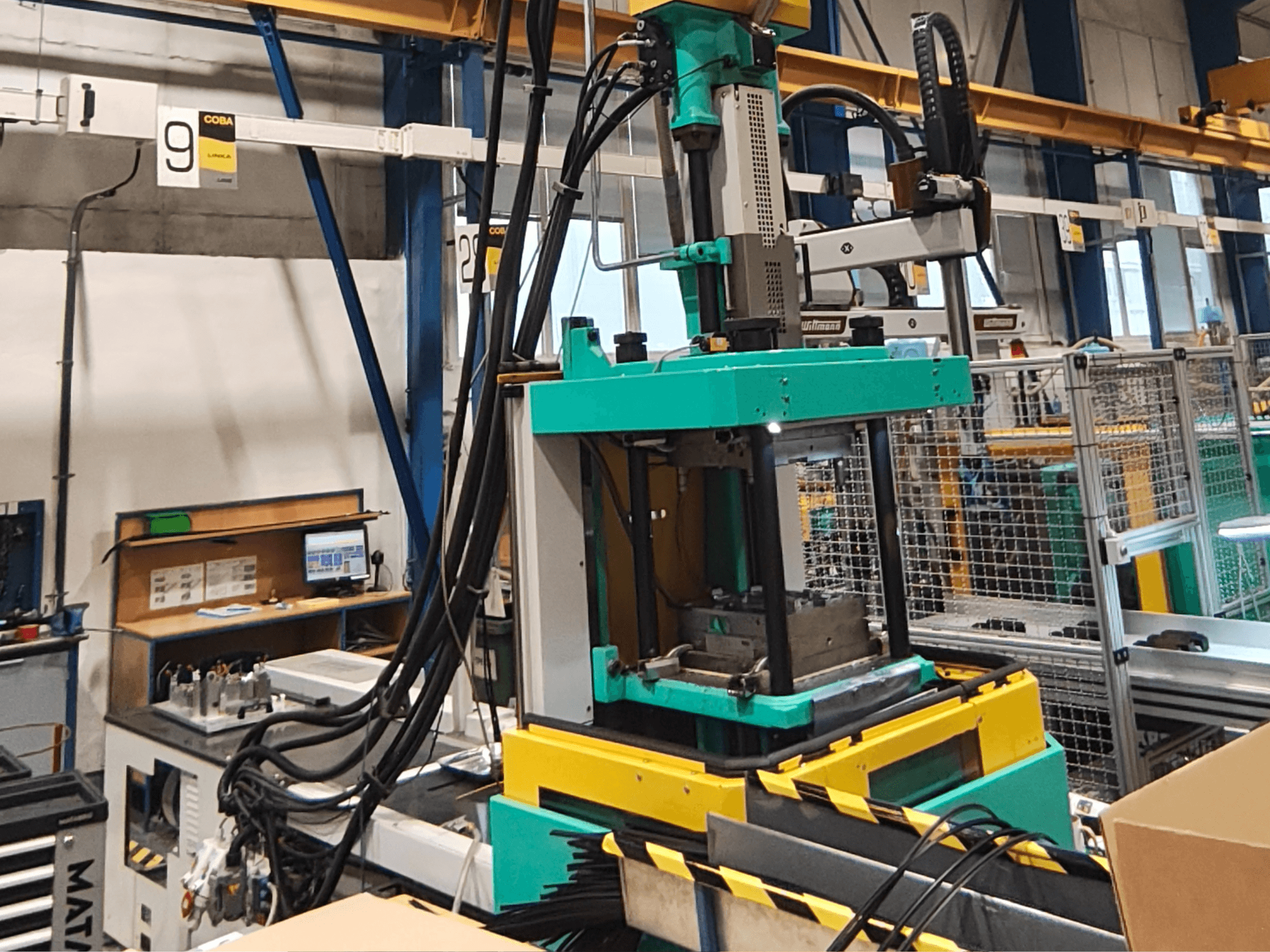 Presse hydraulique verte et jaune dans une usine, vue de face, avec panneau de commande et câbles visibles sur le côté.