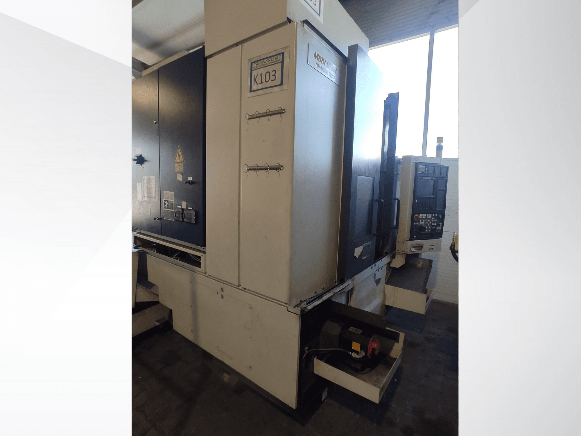 Machine CNC Mori Seiki K103 vue de face, avec panneau de commande, étiquettes de sécurité et composants électriques visibles.