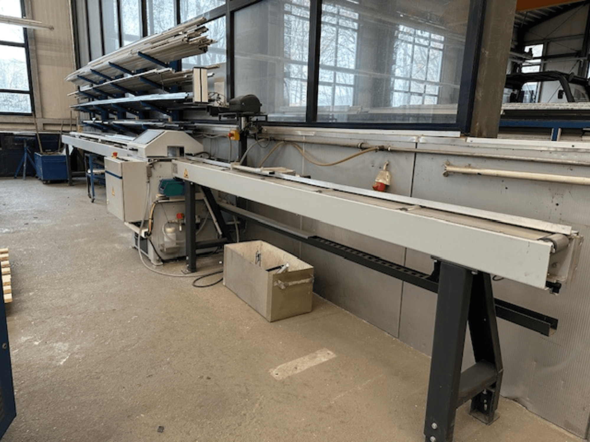 Scie circulaire à métaux SCHÜCO PGS dans un atelier, vue de côté avec un banc de coupe réglable et une étagère de profilés métalliques au-dessus.