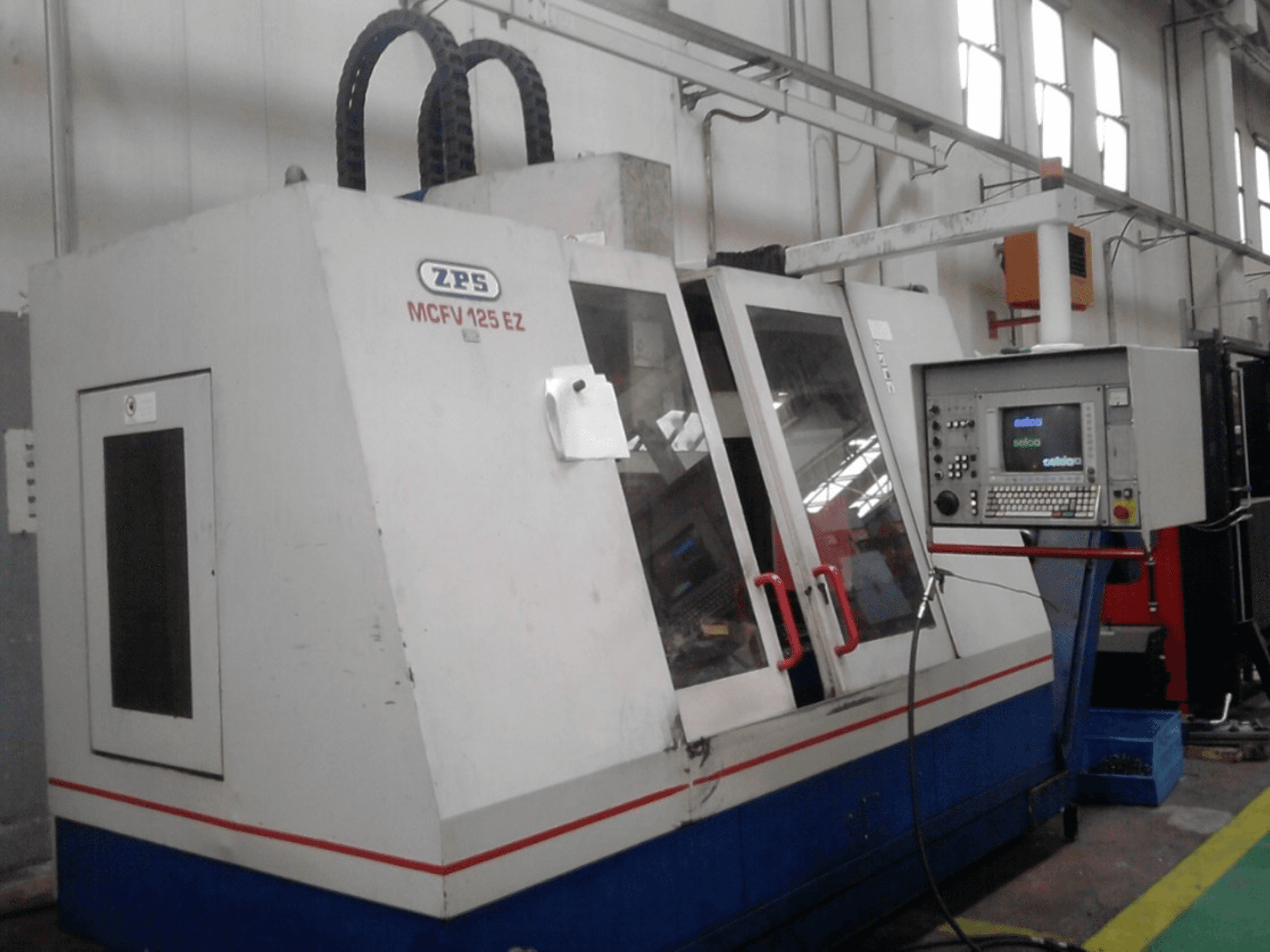 Machine CNC ZPS MCFV 125 EZ, vue de face avec le panneau de commande et les poignées rouges, dans un atelier avec les tuyaux visibles.
