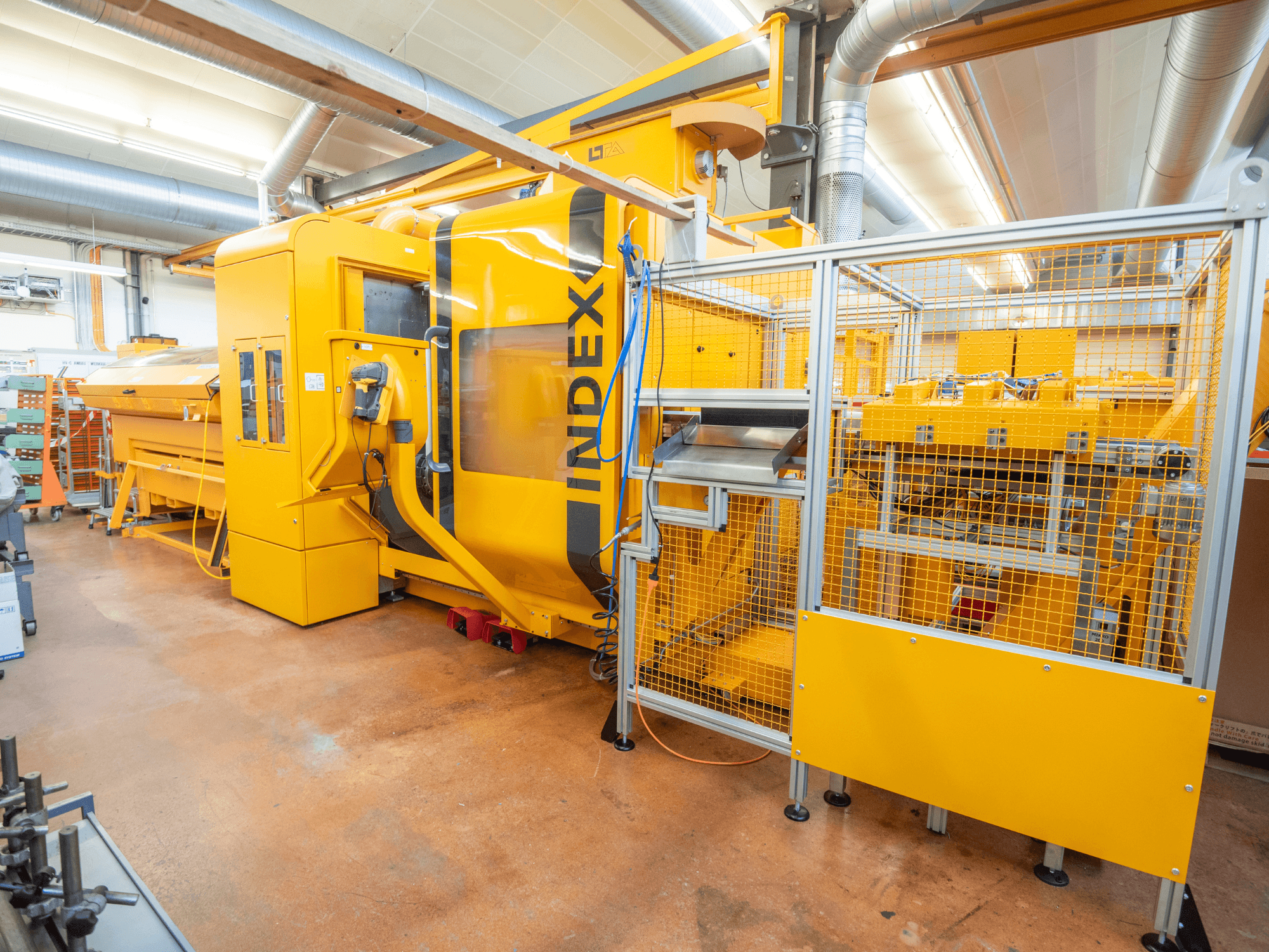Machine INDEX jaune dans un atelier, vue de face. Elle comporte une enceinte de sécurité, un panneau de commande et de nombreux composants opérationnels.