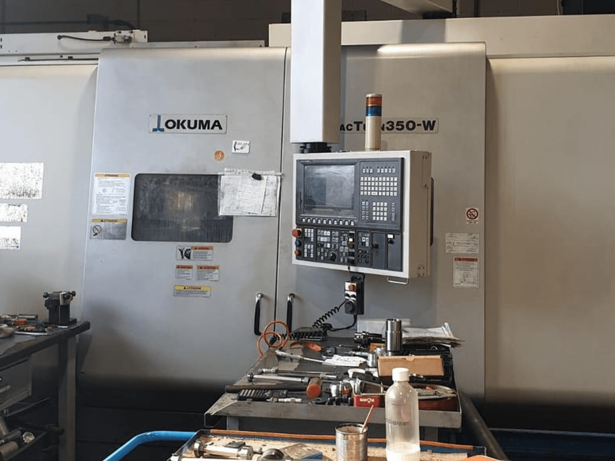 Centre de tournage-fraisage Okuma MAC TURN 350-W, vue de face avec panneau de commande et espace de travail encombré d'outils et d'une bouteille.