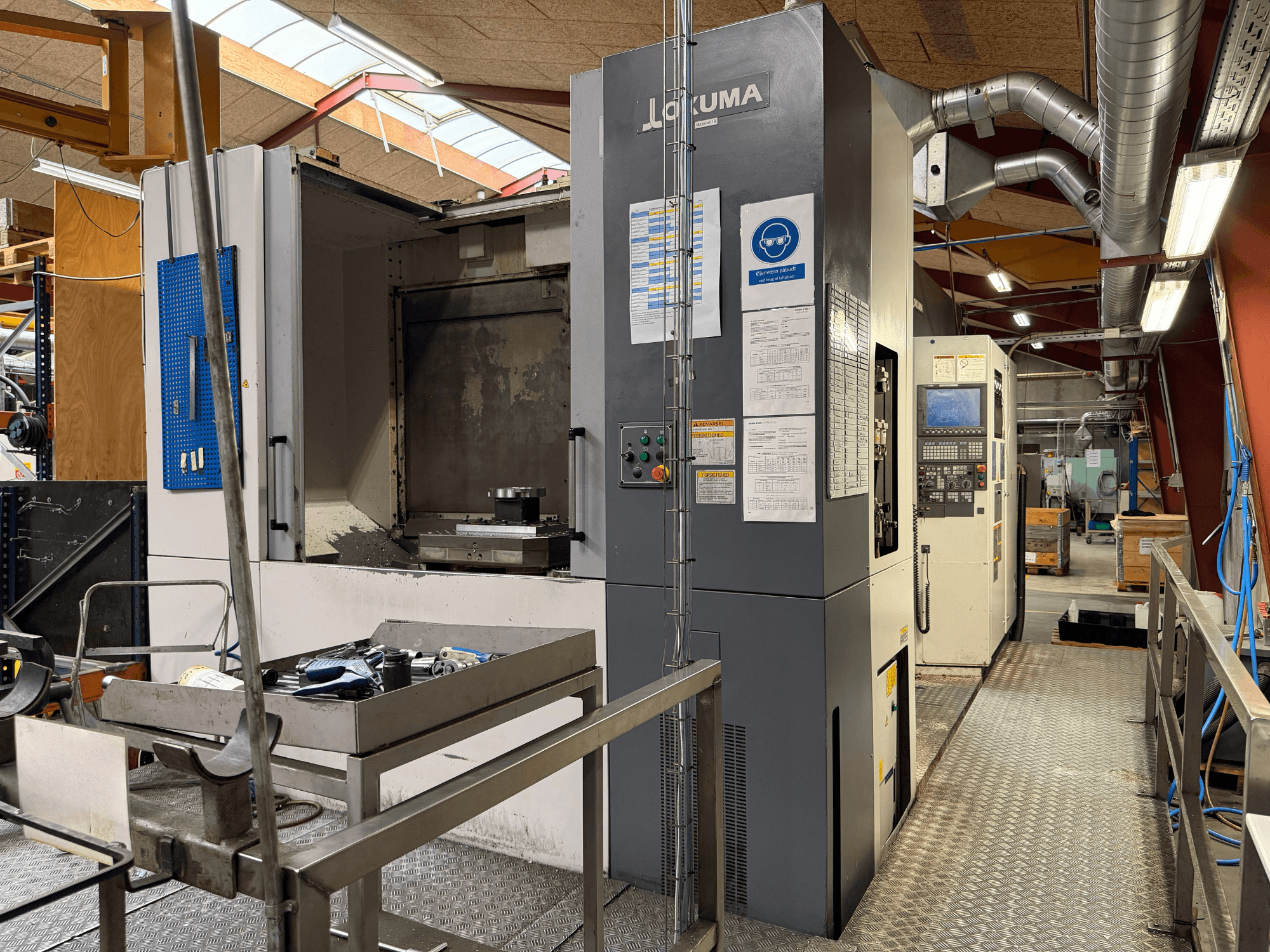 Centre d'usinage horizontal Okuma MA-600HB, vue de face, montrant les outils posés sur une table métallique et les commandes de la machine.