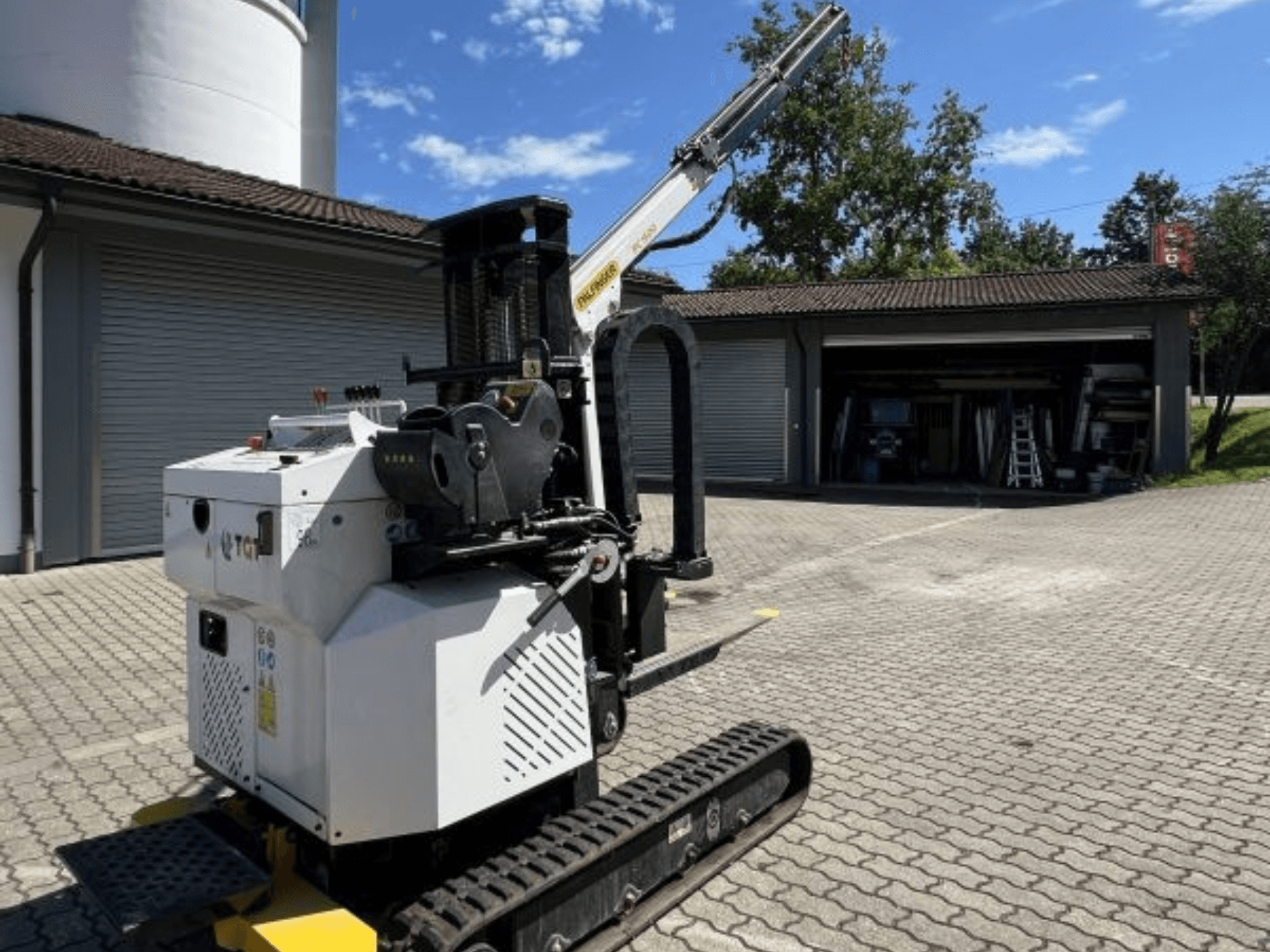 Mini grue de construction TGI, vue latérale, montrant les chenilles et la flèche montée, garée sur une surface en briques près d'un garage.