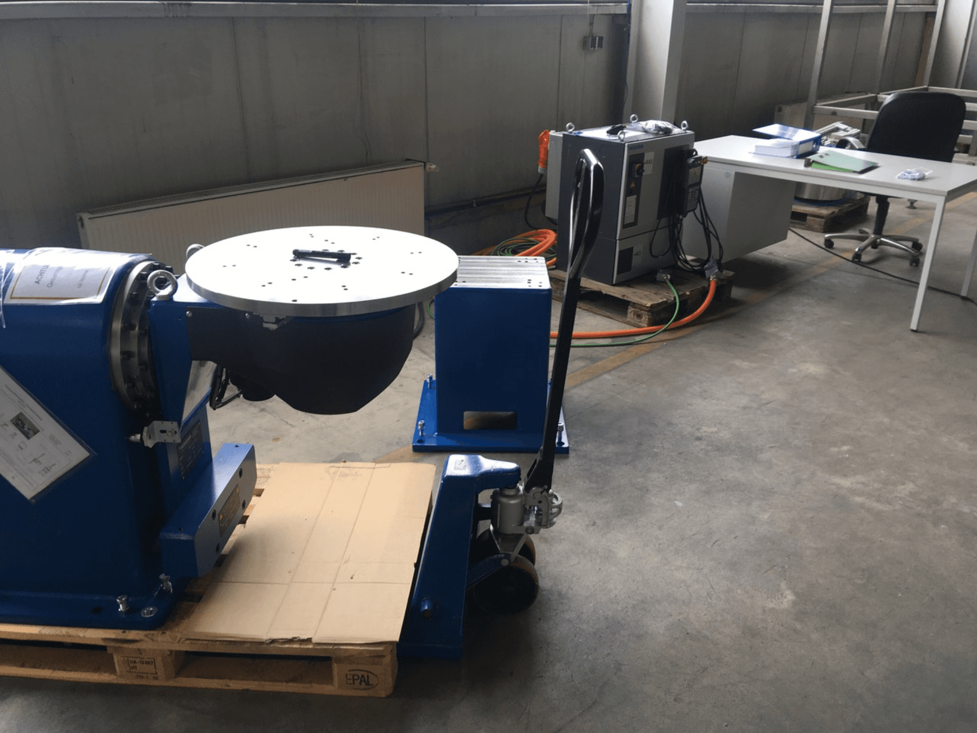 Équipement industriel bleu avec une base circulaire et un panneau de commande, vu de face, sur une palette en bois dans un bureau.