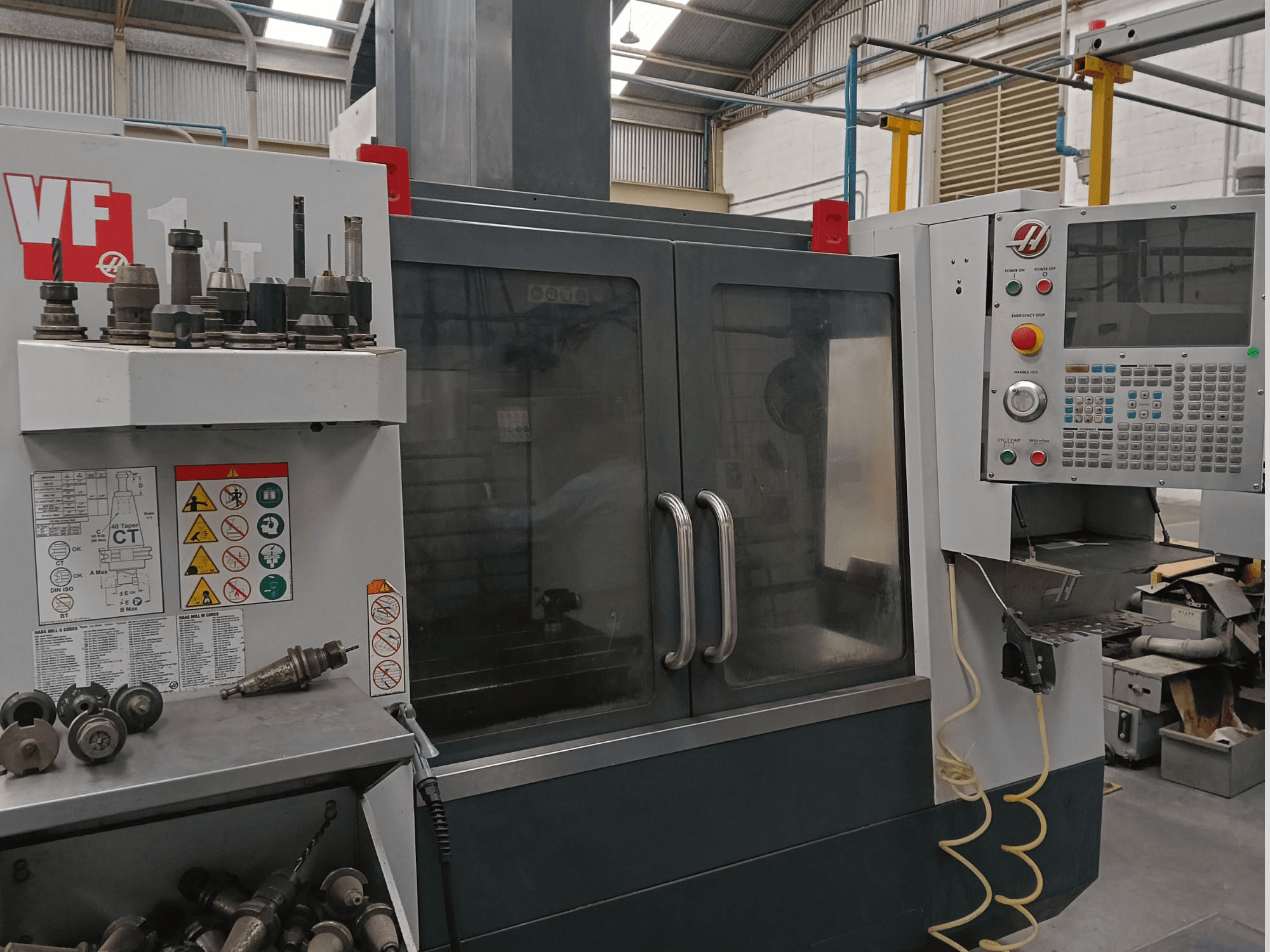 Centre d'usinage vertical HAAS VF-1YT-3 vu de face, mettant en évidence les commandes, les porte-outils et les étiquettes de sécurité visibles.