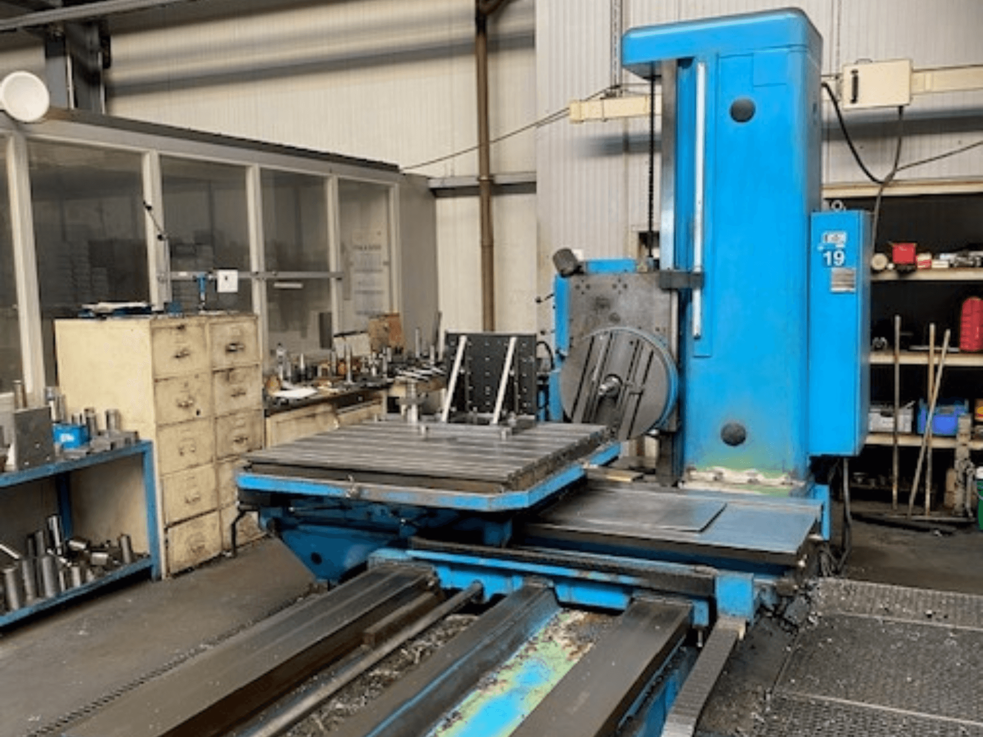 Fraiseuse bleue vue de face, modèle numéro 19, sur le sol d'un atelier avec des outils et une armoire de rangement à l'arrière-plan.