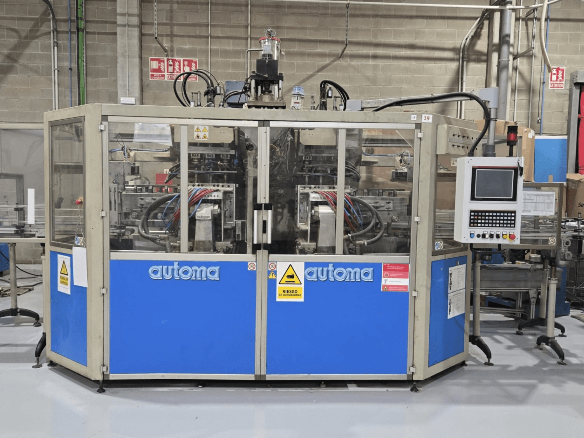 Vue de face de la machine de moulage par soufflage AUTOMA APEX AT500D, avec son panneau de commande et ses différents tuyaux, installée dans un environnement industriel.