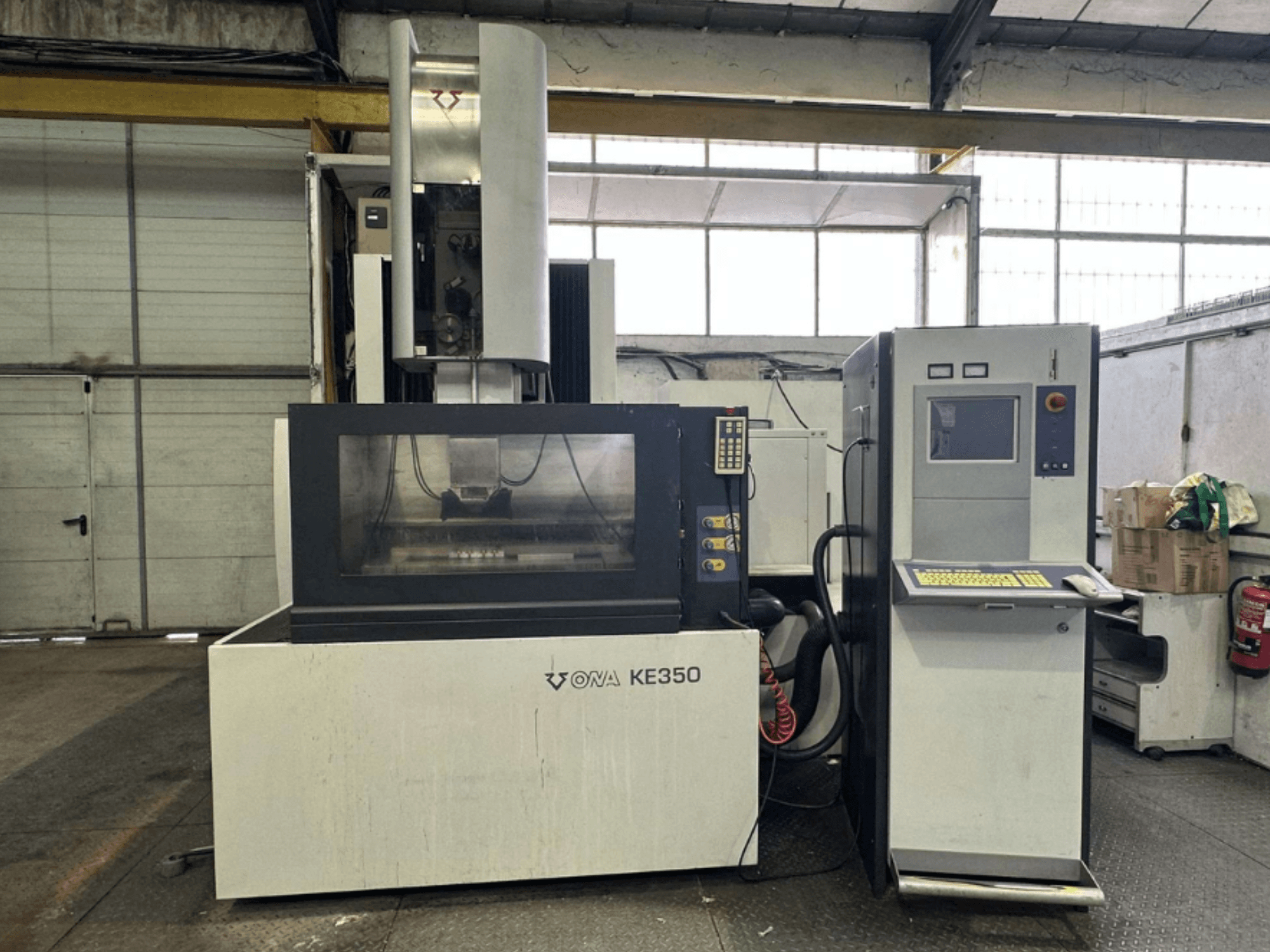 Vue frontale de la machine CNC ONA KE350 avec panneau de commande, écran d'affichage et divers boutons, située dans un environnement industriel.