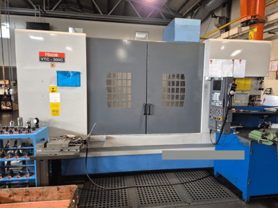 Machine Mazak VTC-300C dans une usine, vue de face montrant le panneau de commande, les portes grises avec la grille des fenêtres, et l'équipement à proximité.