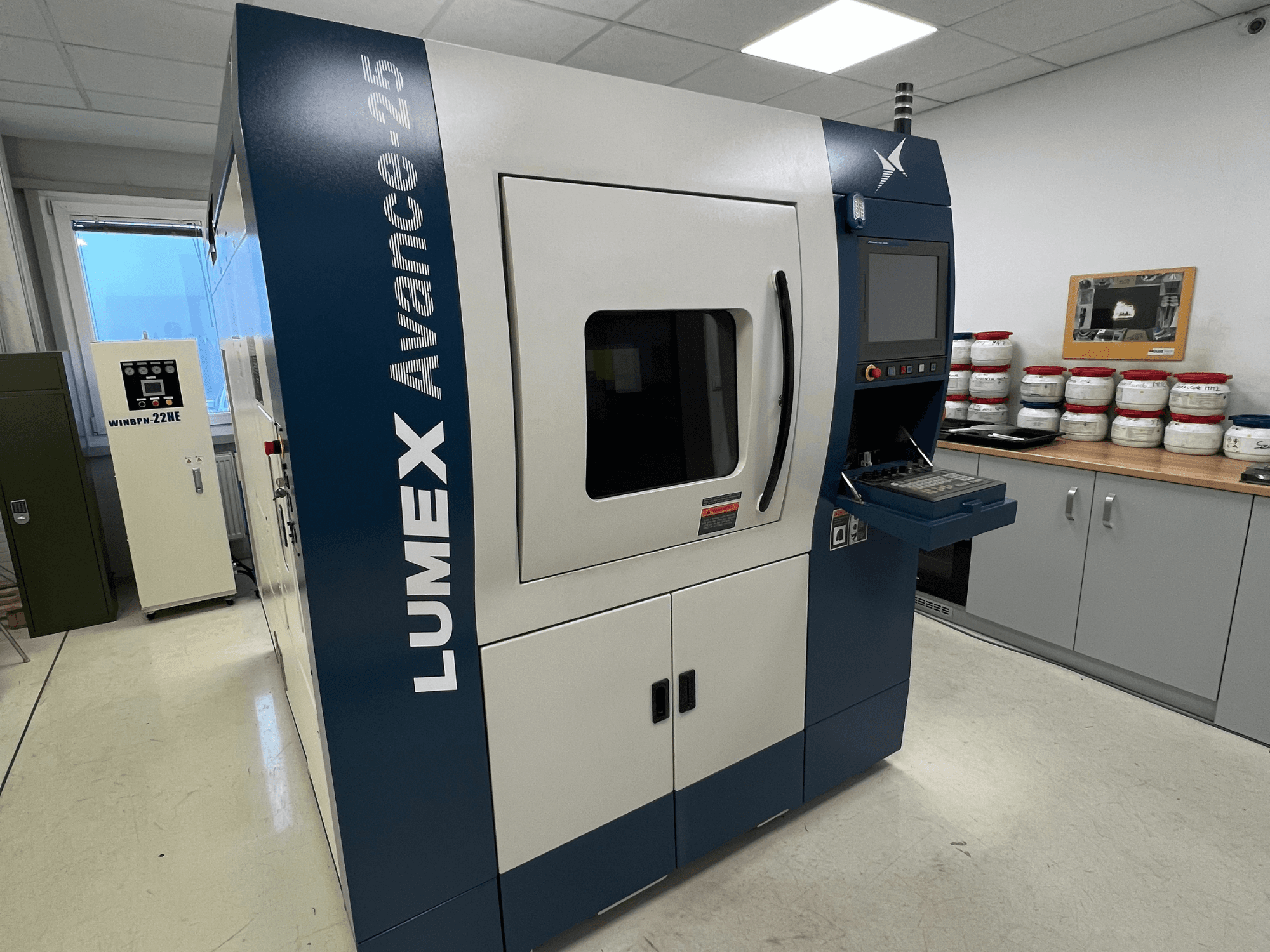 Imprimante 3D LUMEX Avance-25 dans un laboratoire, vue de face. Panneau de commande avec écran et boutons visibles, équipement en arrière-plan.