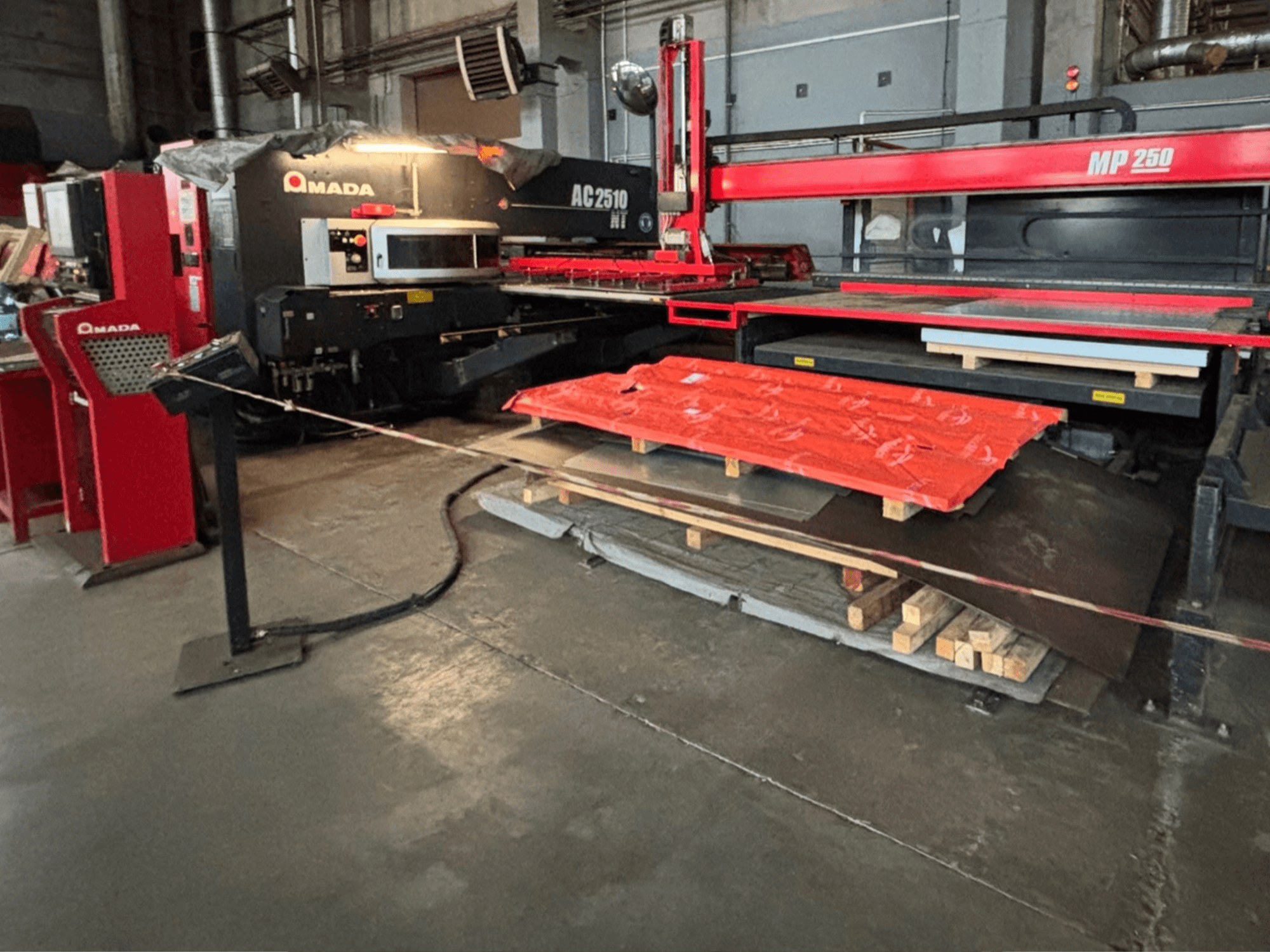 Découpeur laser Amada AC2510 NT en usine, vue de face, machines lourdes avec tôles et palettes au sol.