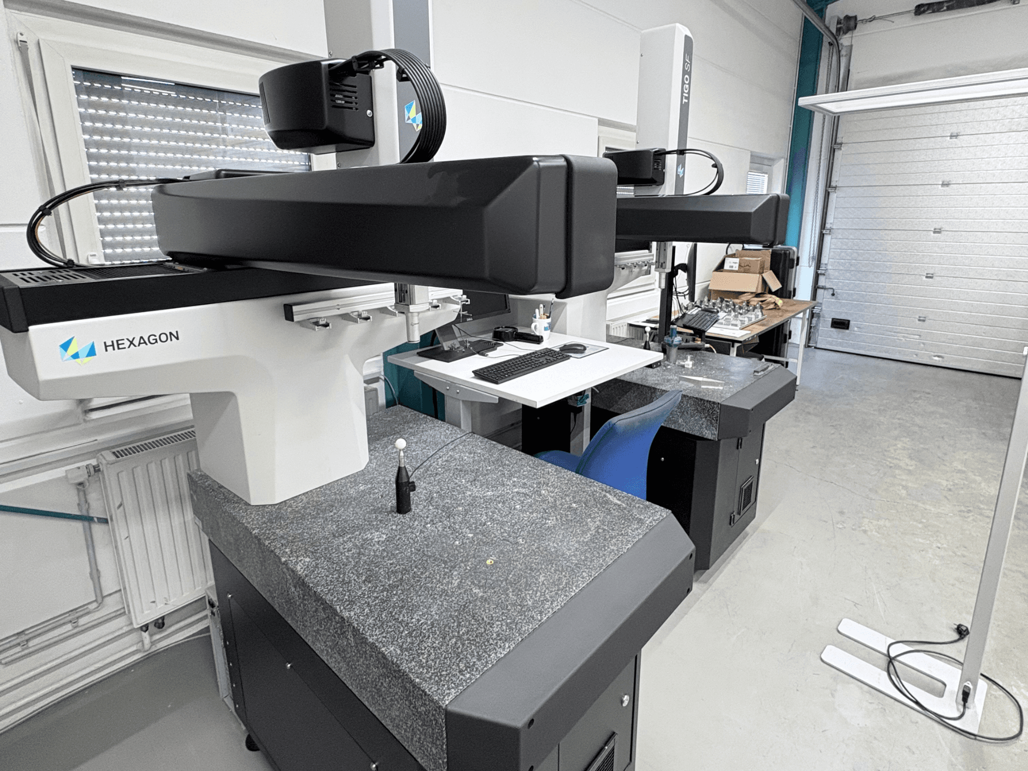 Machine à mesurer tridimensionnelle (CMM) Hexagon Tigo SF dans l'atelier, équipée d'un socle en granit, d'une sonde de mesure et d'un poste de travail à proximité avec des outils.