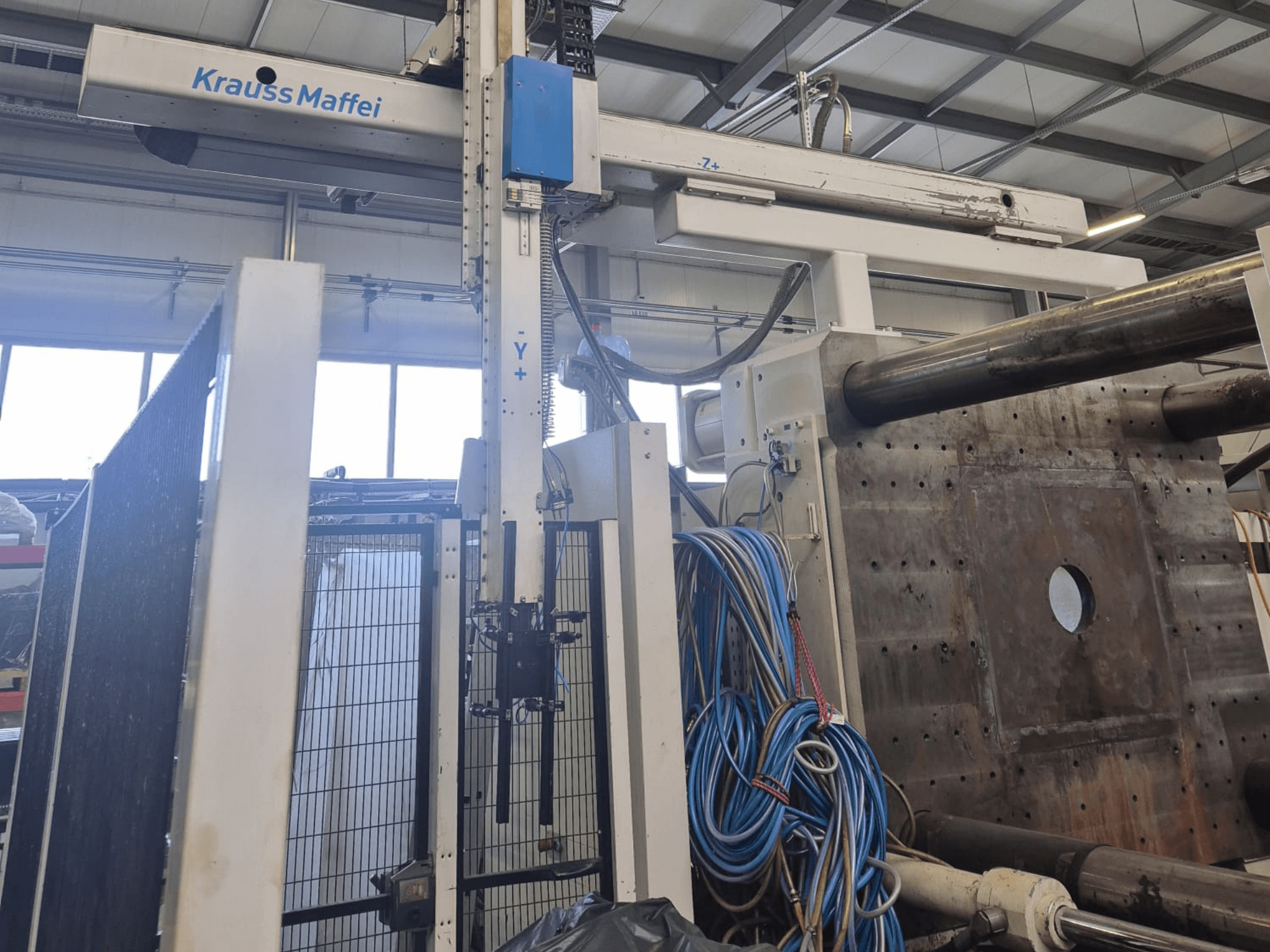 Presse à injection hydraulique Krauss Maffei 1300-12000 MX, vue de face avec bras mécaniques et raccords hydrauliques.