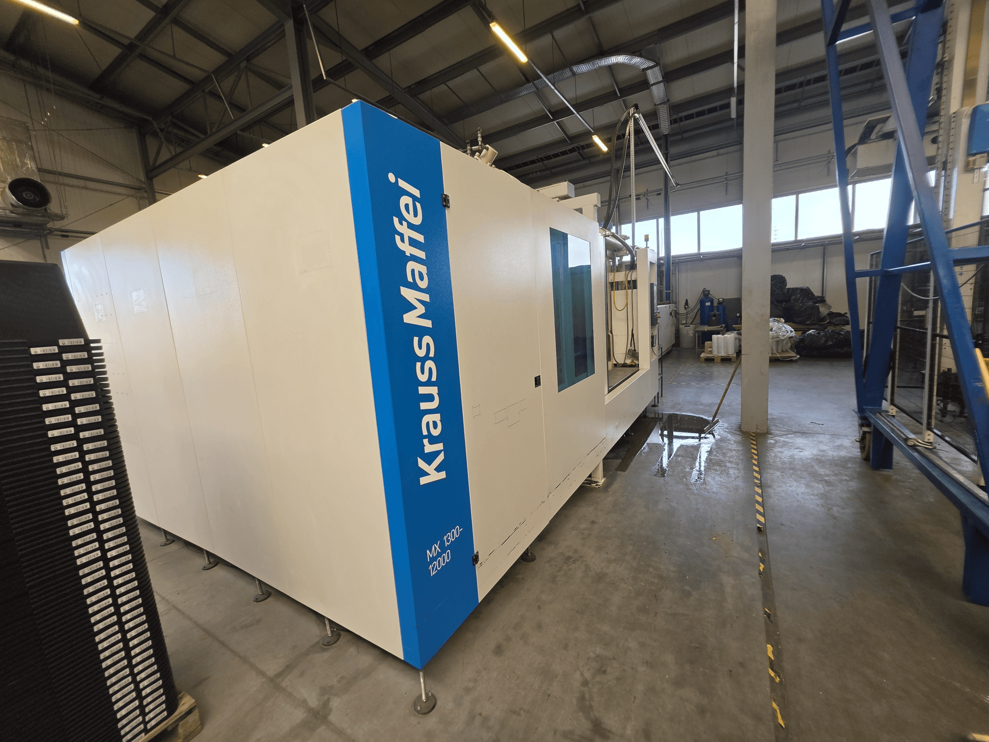 Machine de moulage par injection hydraulique Krauss Maffei MX 1300-12000 vue de profil, mettant en valeur son grand châssis blanc et son logo bleu.