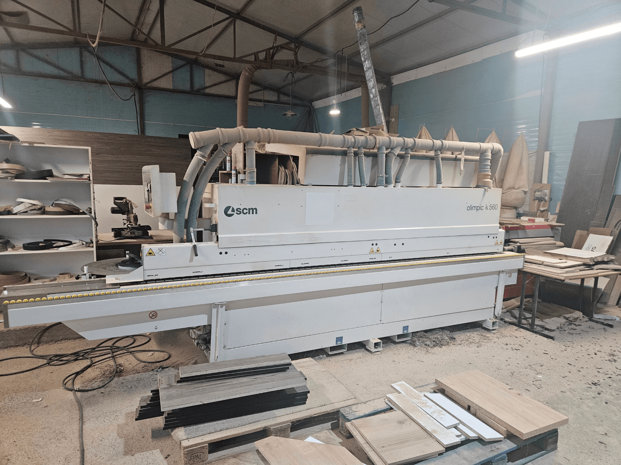 Plaqueuse de chants SCM Olimpic K560 dans un atelier, vue de face, avec des panneaux de bois et des outils visibles sur le sol.