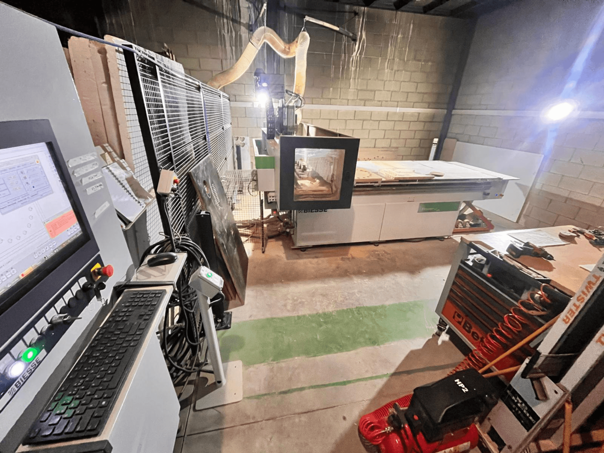 Machine CNC Biesse dans un atelier, vue latérale montrant le panneau de commande, l'aspiration des poussières, les outils et divers matériaux sur la table.