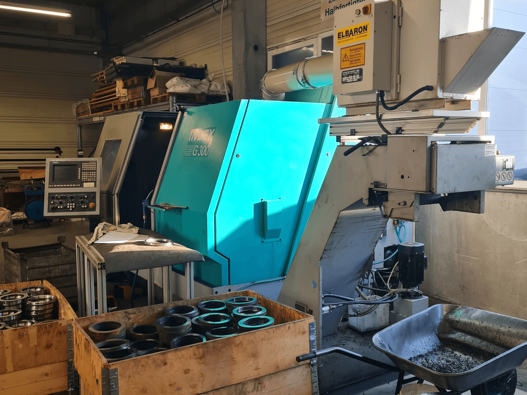 Vue de face du tour horizontal Index G300 RatioLine avec son panneau de commande, entouré de pièces et d'un convoyeur.