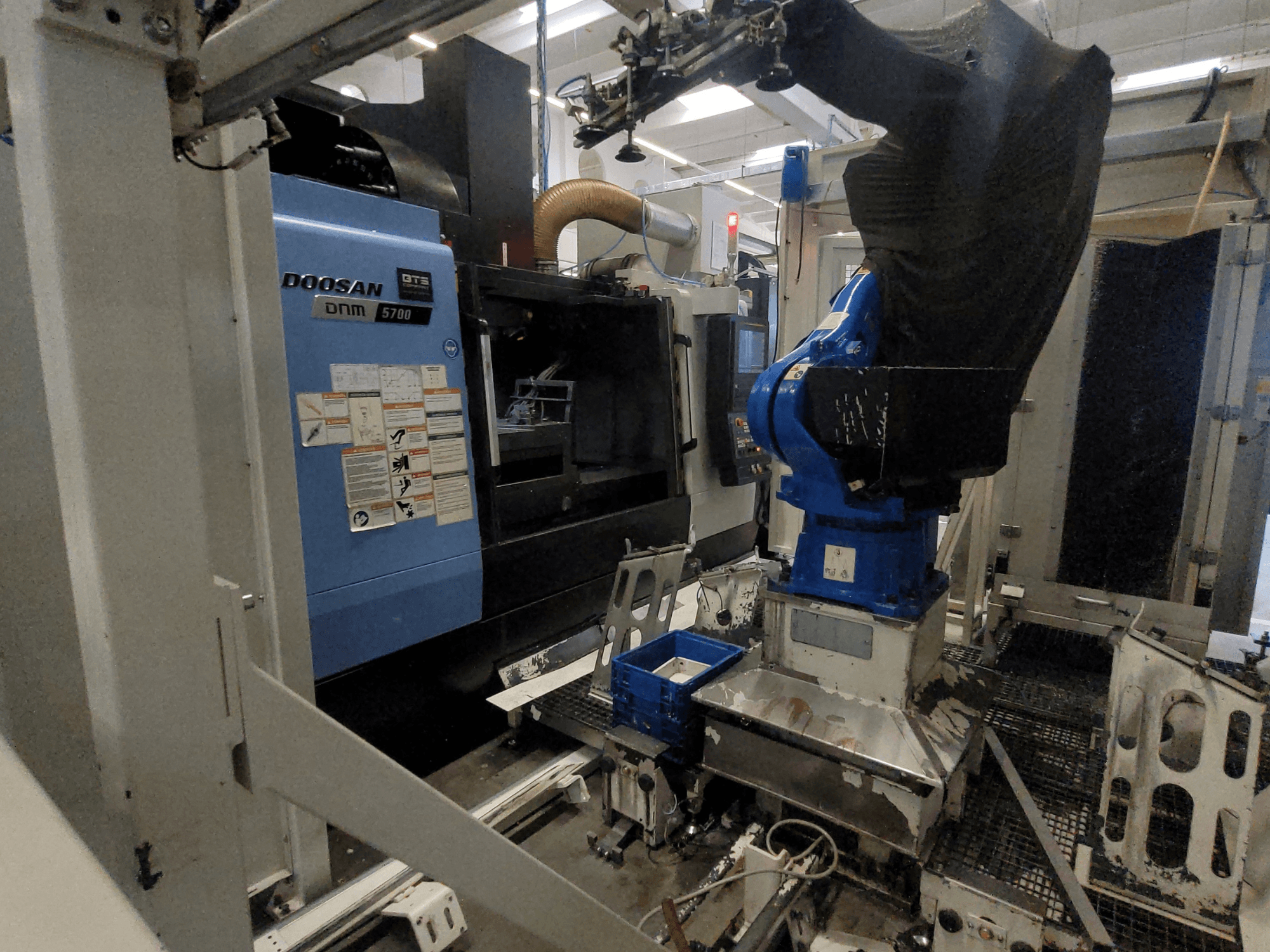 Centre d'usinage vertical DOOSAN DNM 5700 dans un environnement d'usine, présentant sa vue de face et l'intégration d'un bras robotisé.