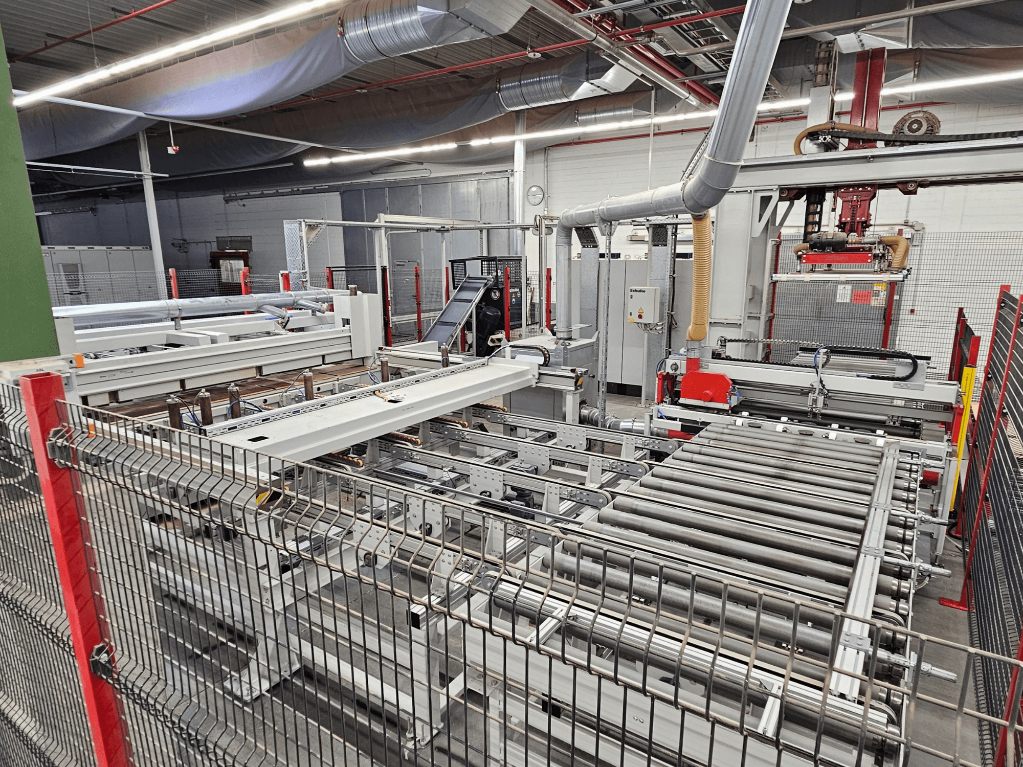 La scie à panneaux HOLZMA HPP81-3200 est présentée en vue de face, avec des rouleaux de transport et des composants de machines dans un atelier.