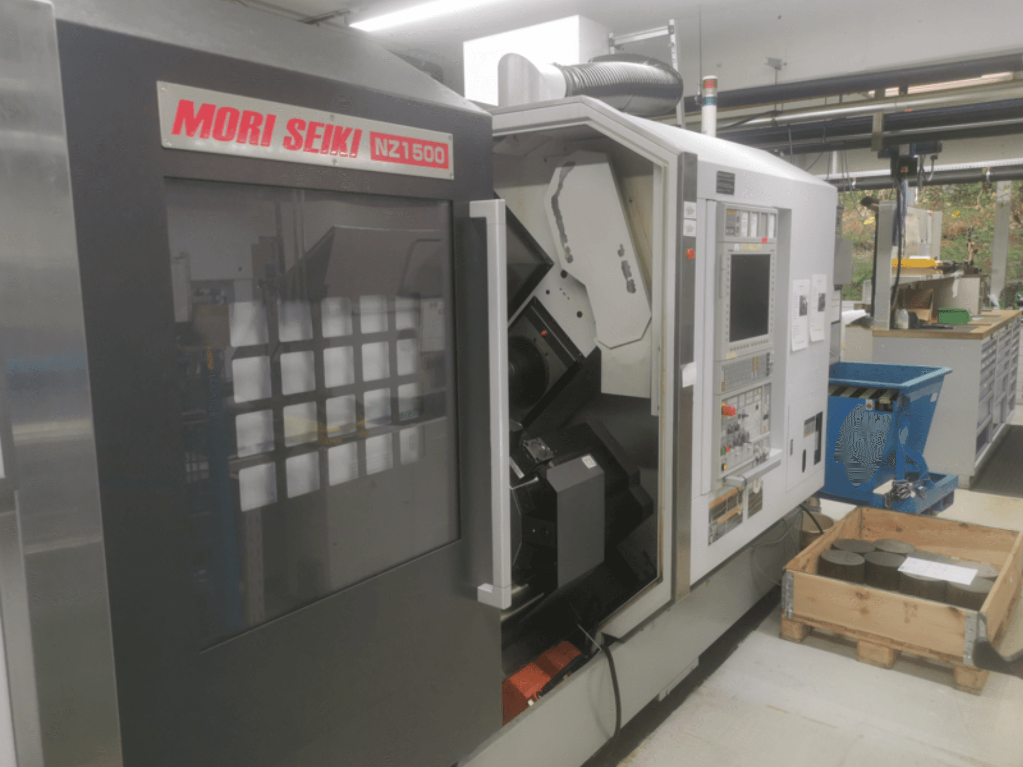 Machine CNC MORI SEIKI NZ1500 légèrement inclinée, montrant le panneau de commande et l'espace de travail avec des pièces métalliques sur une palette en bois.