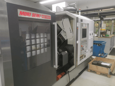 Machine CNC MORI SEIKI NZ1500 légèrement inclinée, montrant le panneau de commande et l'espace de travail avec des pièces métalliques sur une palette en bois.