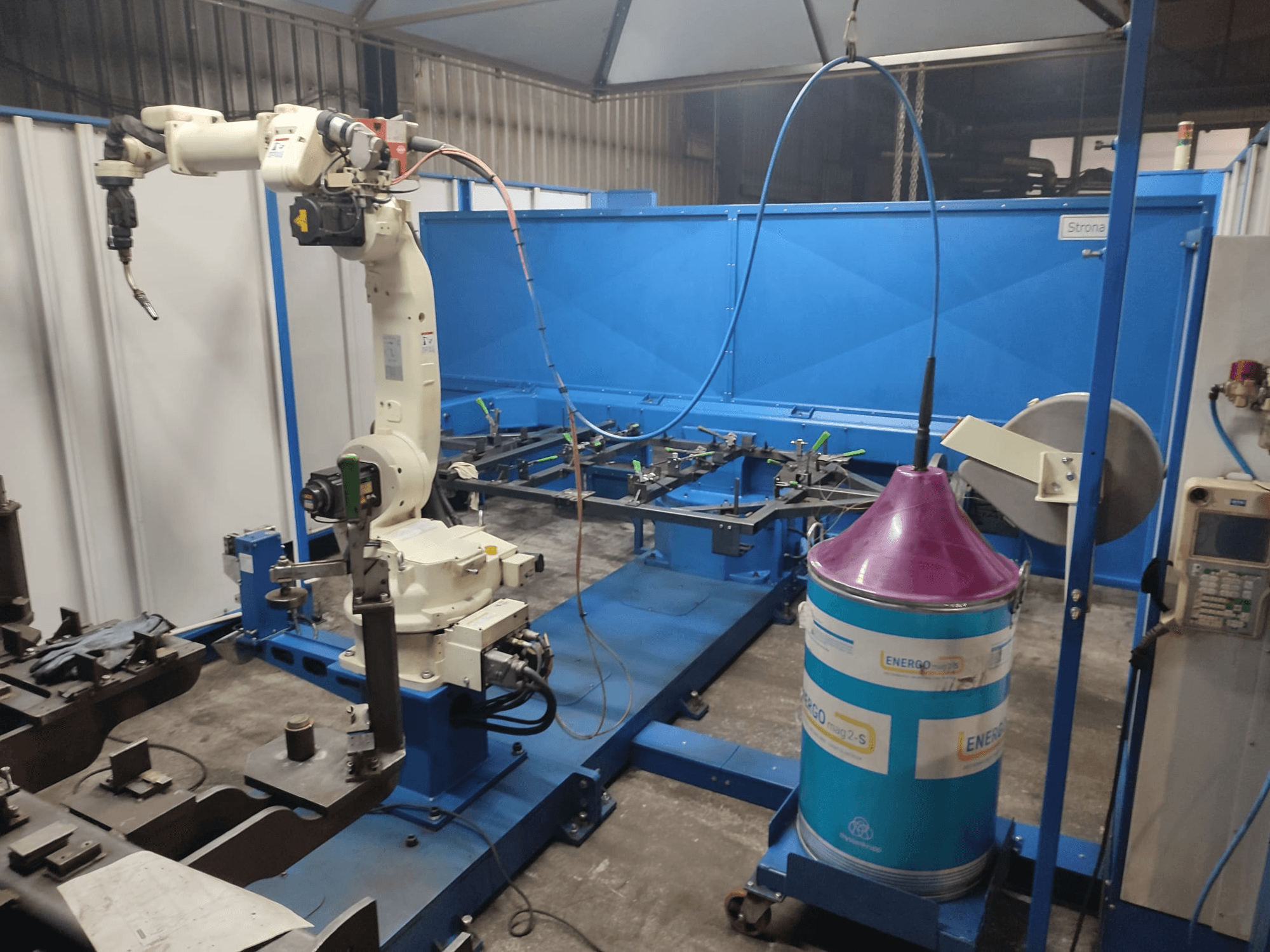 Bras robotique industriel avec une base blanche et bleue, installé dans un atelier avec un plan de travail bleu et des composants métalliques.