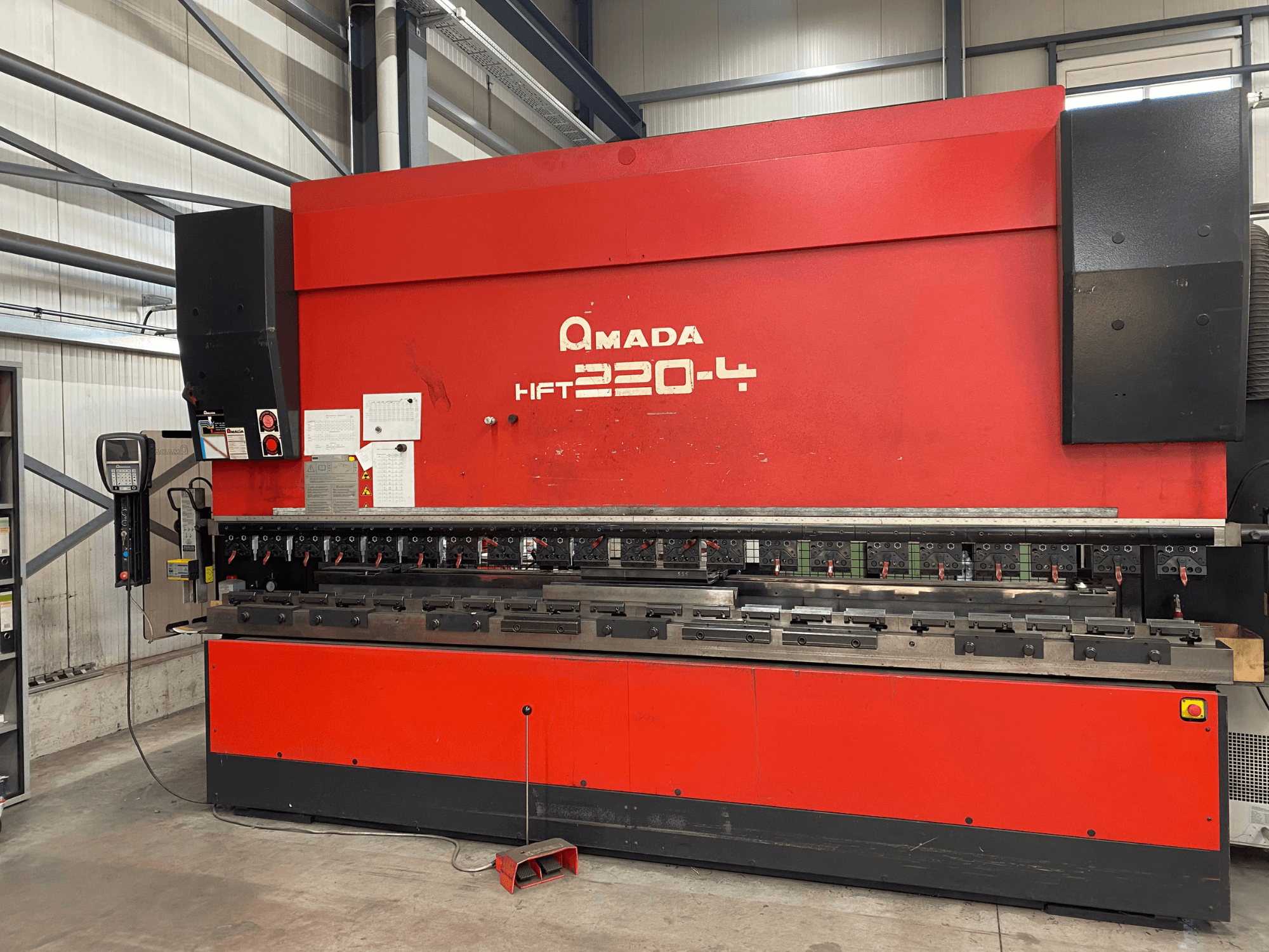 Presse plieuse AMADA HFT 220-4 rouge en usine, vue de face montrant le panneau de commande et les outils de pliage sur la surface de travail.