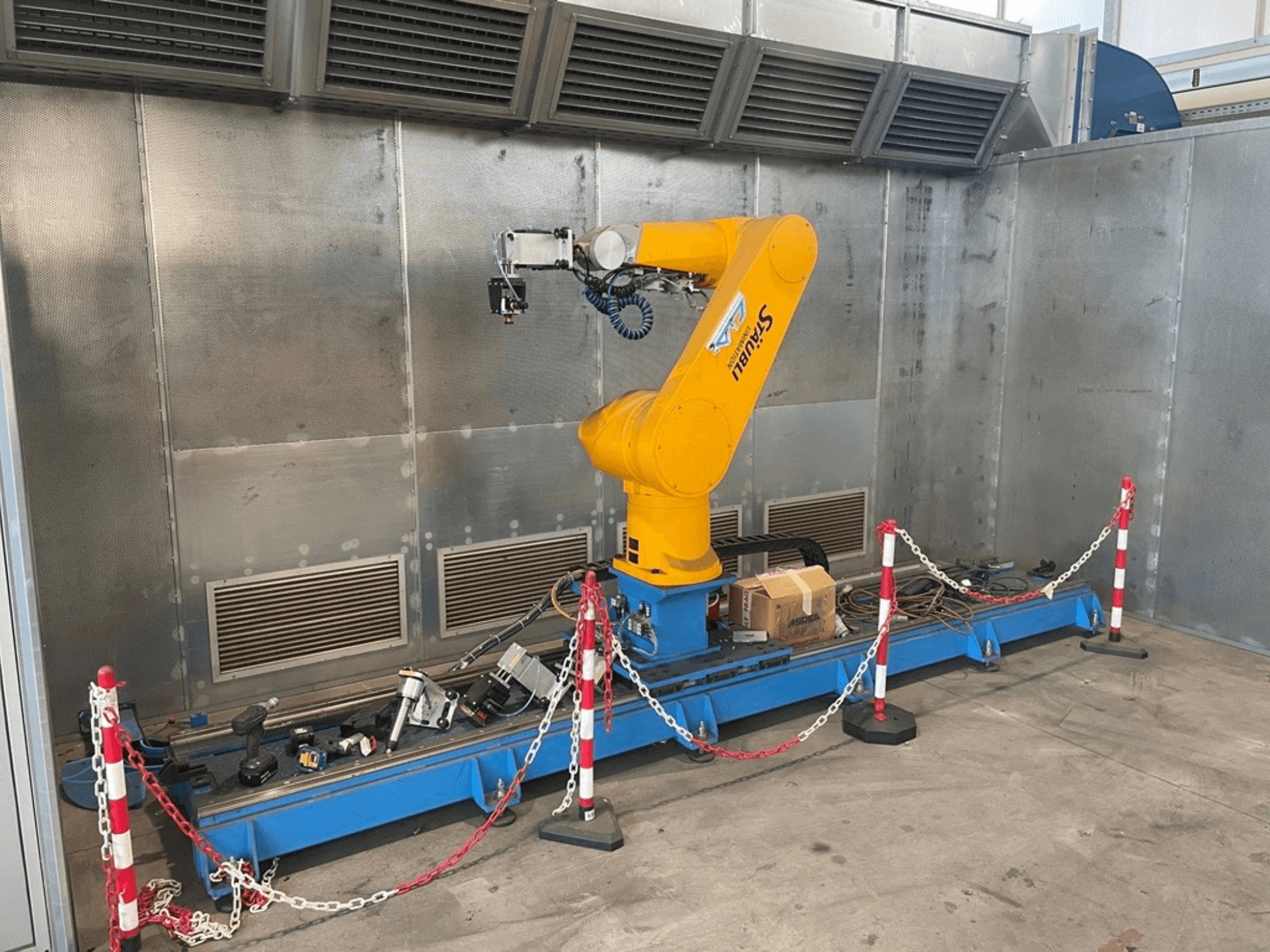 Bras robotique STAUBLI CS8C-RX160 dans un atelier, vue latérale, avec des câbles et divers outils sur une base bleue entourée de barrières de protection.