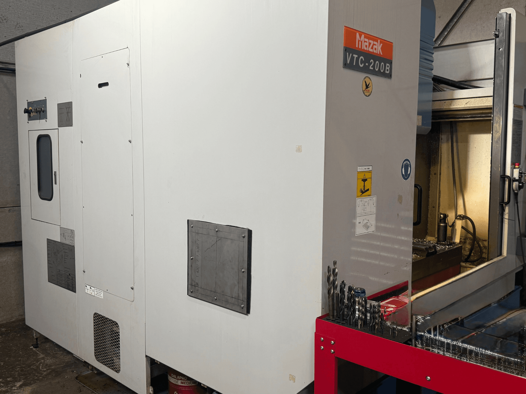Centre d'usinage vertical Mazak VTC-200B vu de profil, avec les commandes identifiées et les forets rangés sur un support de rangement rouge.