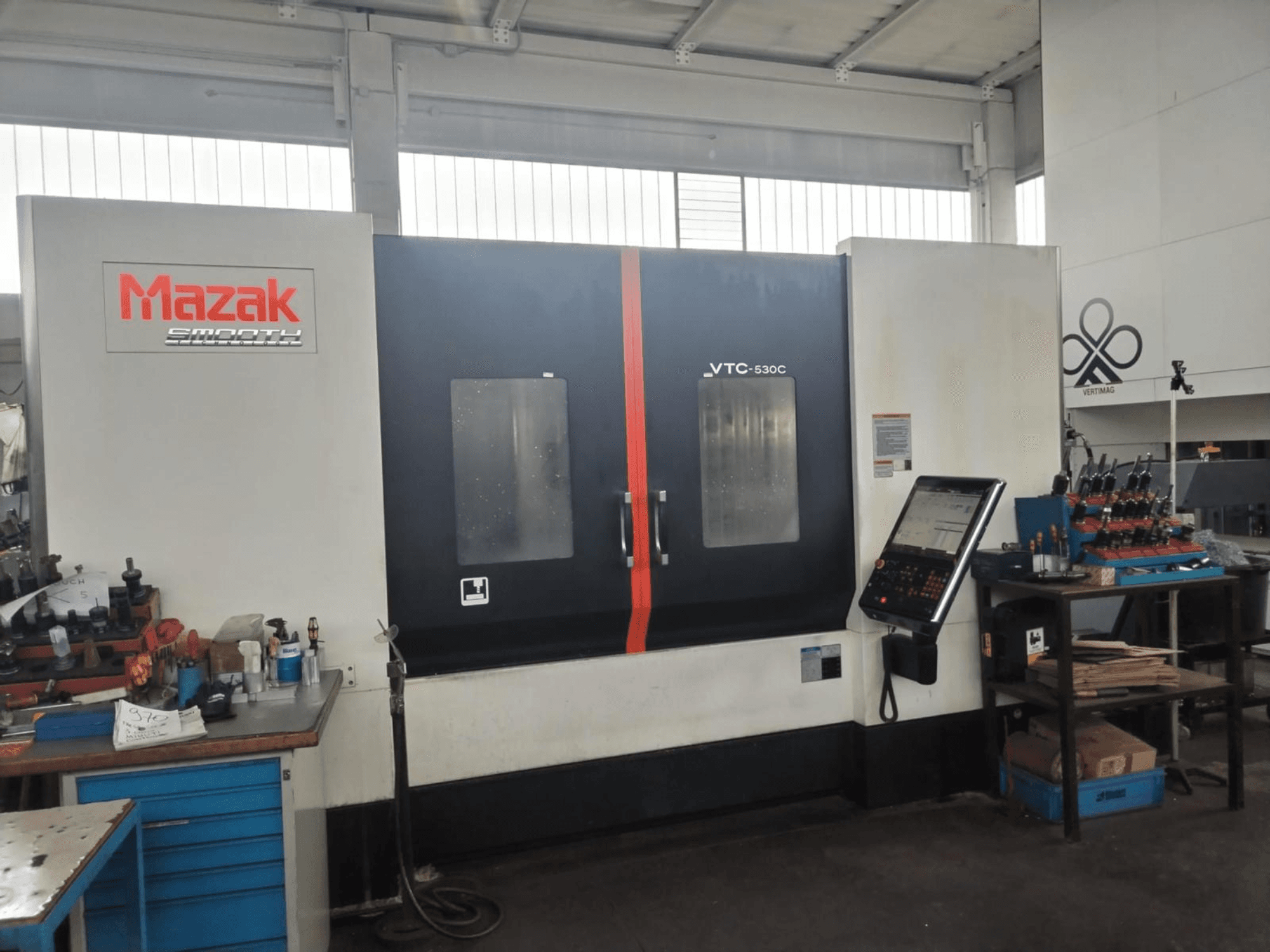 Centre d'usinage vertical Mazak VTC-530C dans un atelier, avec un panneau de commande et des outils posés sur une table d'appoint.