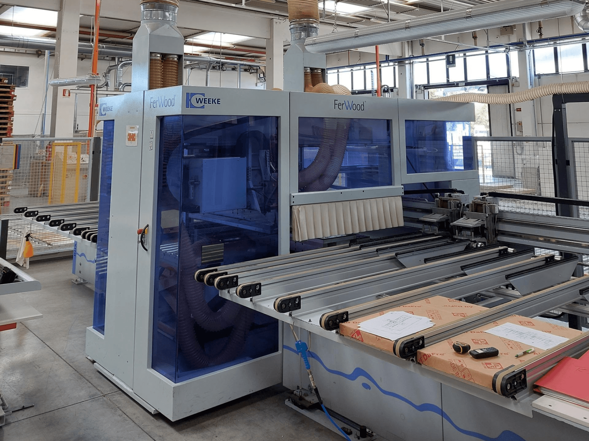 Machine CNC Weeke FerWood dans un atelier, vue de face montrant les panneaux teintés en bleu, les boutons de commande et la table de travail avec les matériaux.